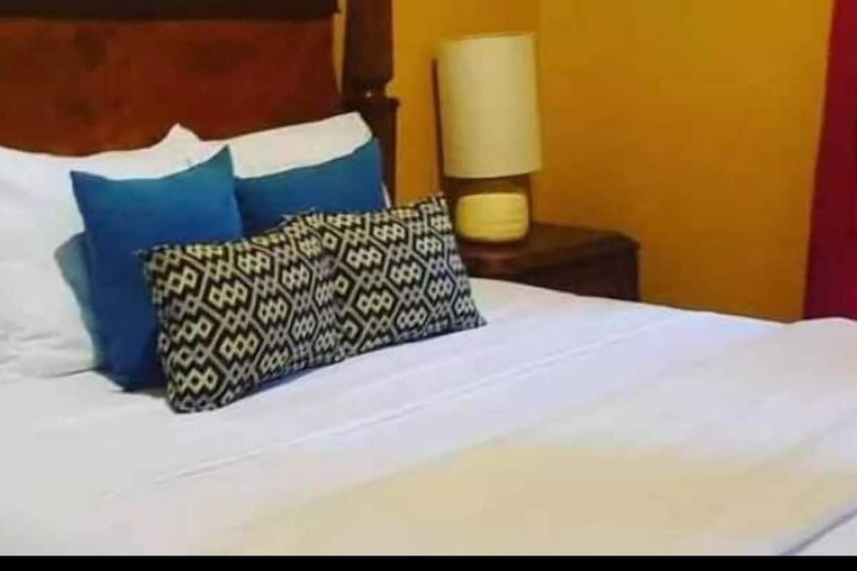 A bed with white sheets, two blue pillows, and a patterned pillow. There is a wooden headboard, a bedside table with a lamp, and a wall clock.
