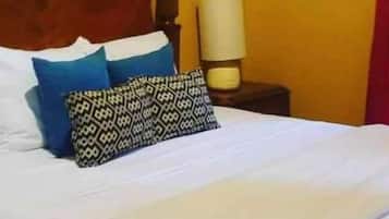 Una cama con sábanas blancas, dos almohadas azules y una almohada estampada. Tiene un cabecero de madera, una mesita de noche con una lámpara y un reloj de pared.