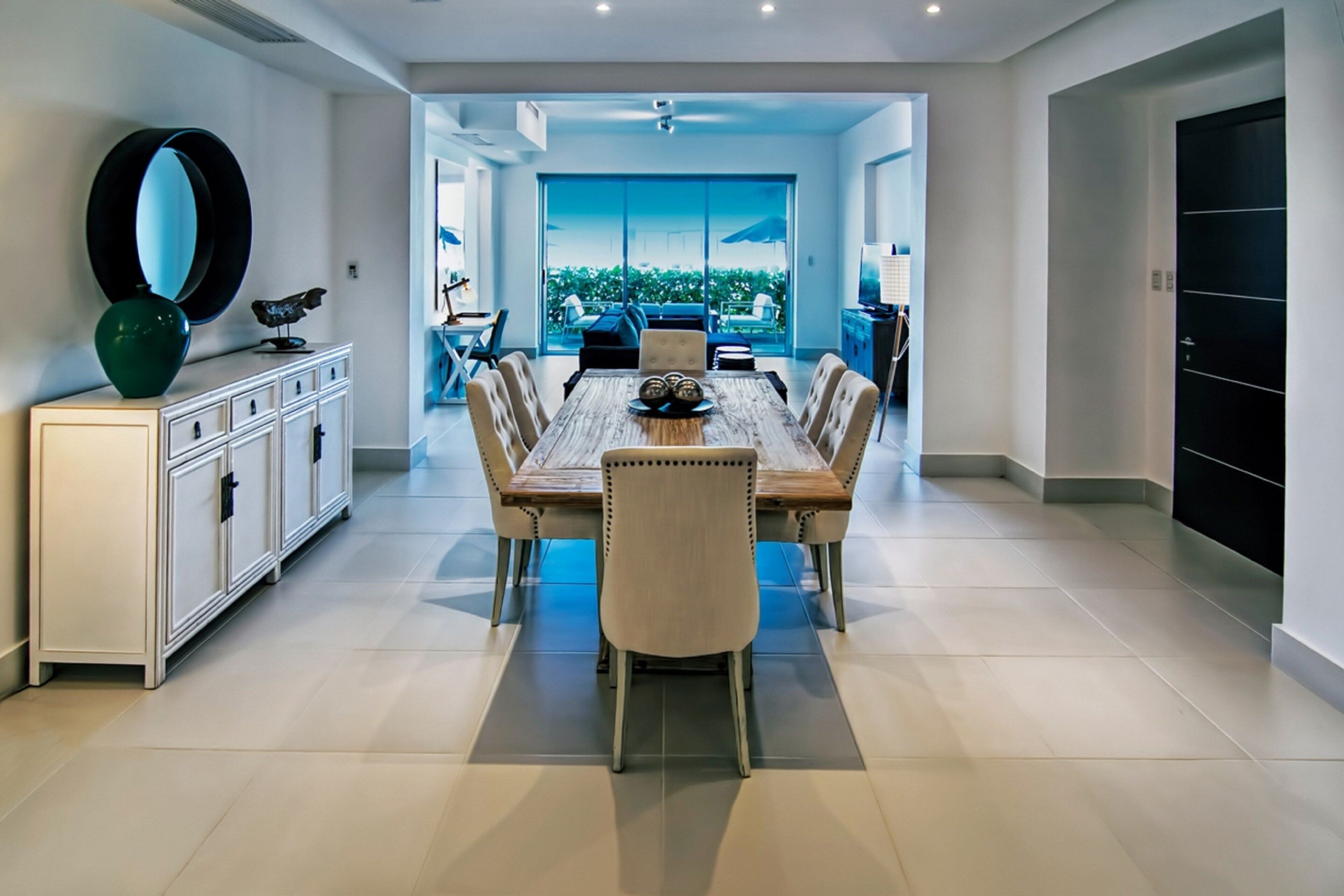 A spacious dining area with a long wooden table and six chairs, leading to a balcony with a view of greenery.
