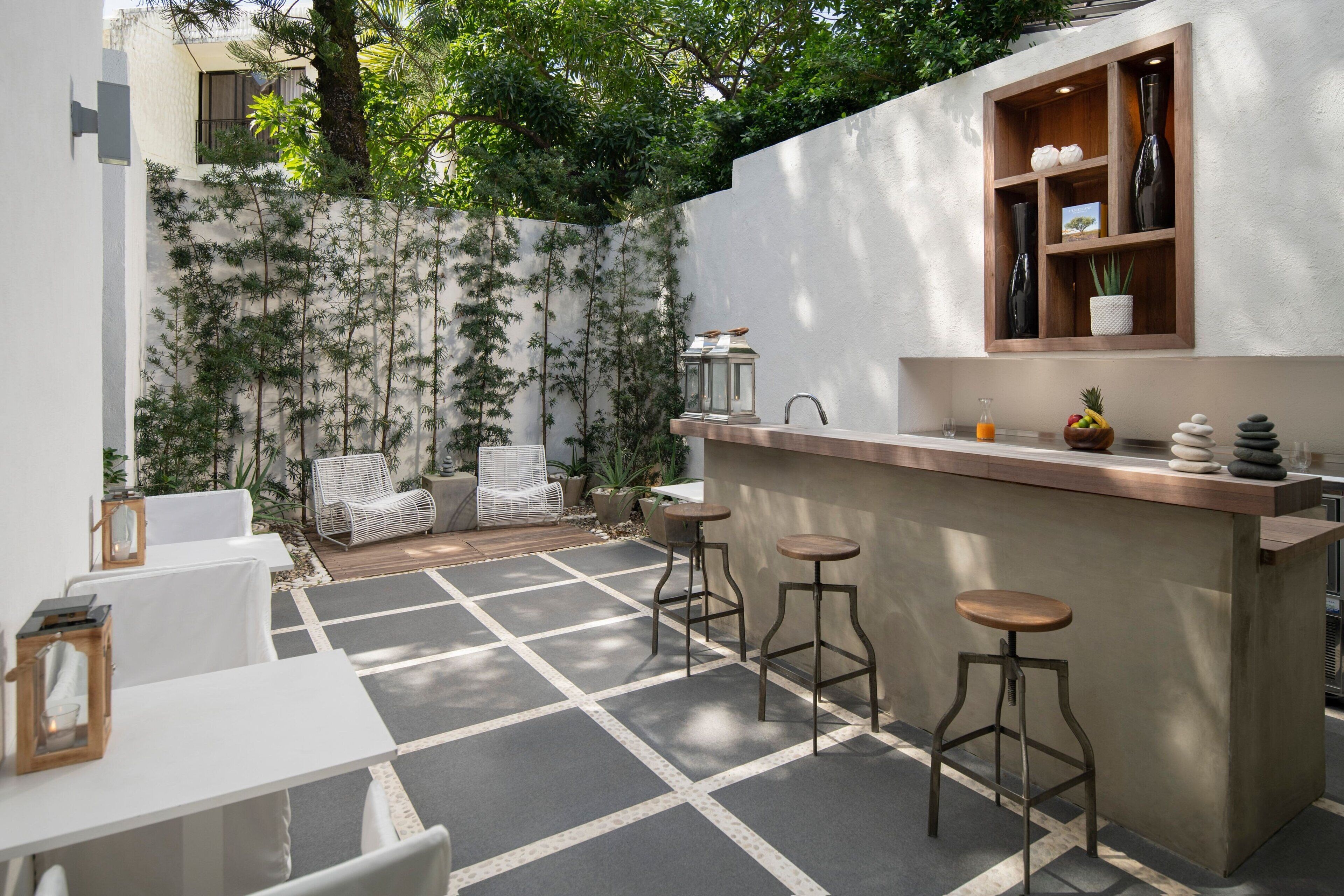 Un moderno espacio al aire libre con barra, suelo de baldosas blancas y grises, mostrador de madera y tres taburetes. Se observan sillas blancas y una pequeña mesa con un espejo y un farol.