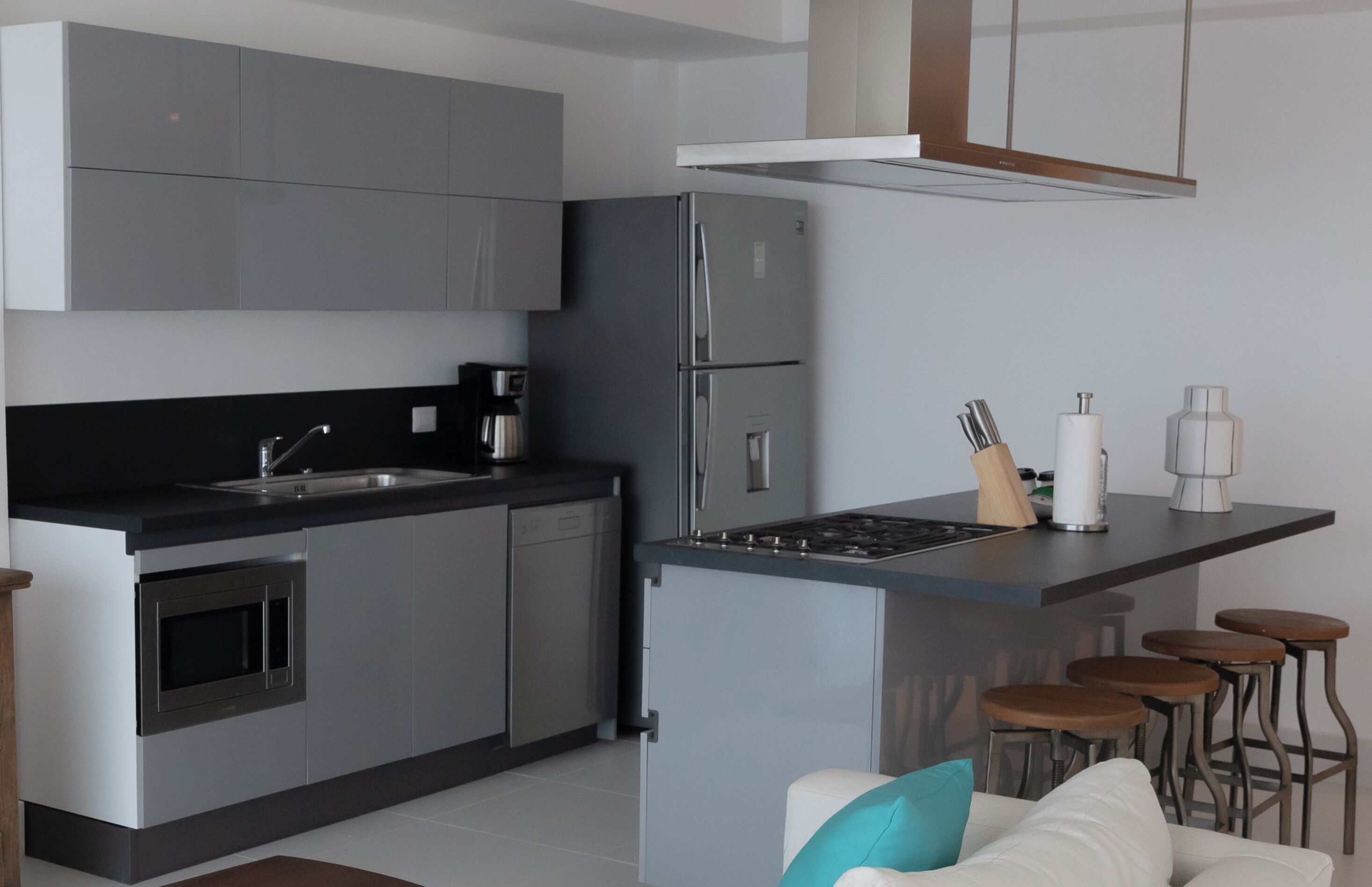 Una cocina moderna con gabinetes grises, un refrigerador de acero inoxidable, horno empotrado, un lavamanos y una zona de bar con taburetes.