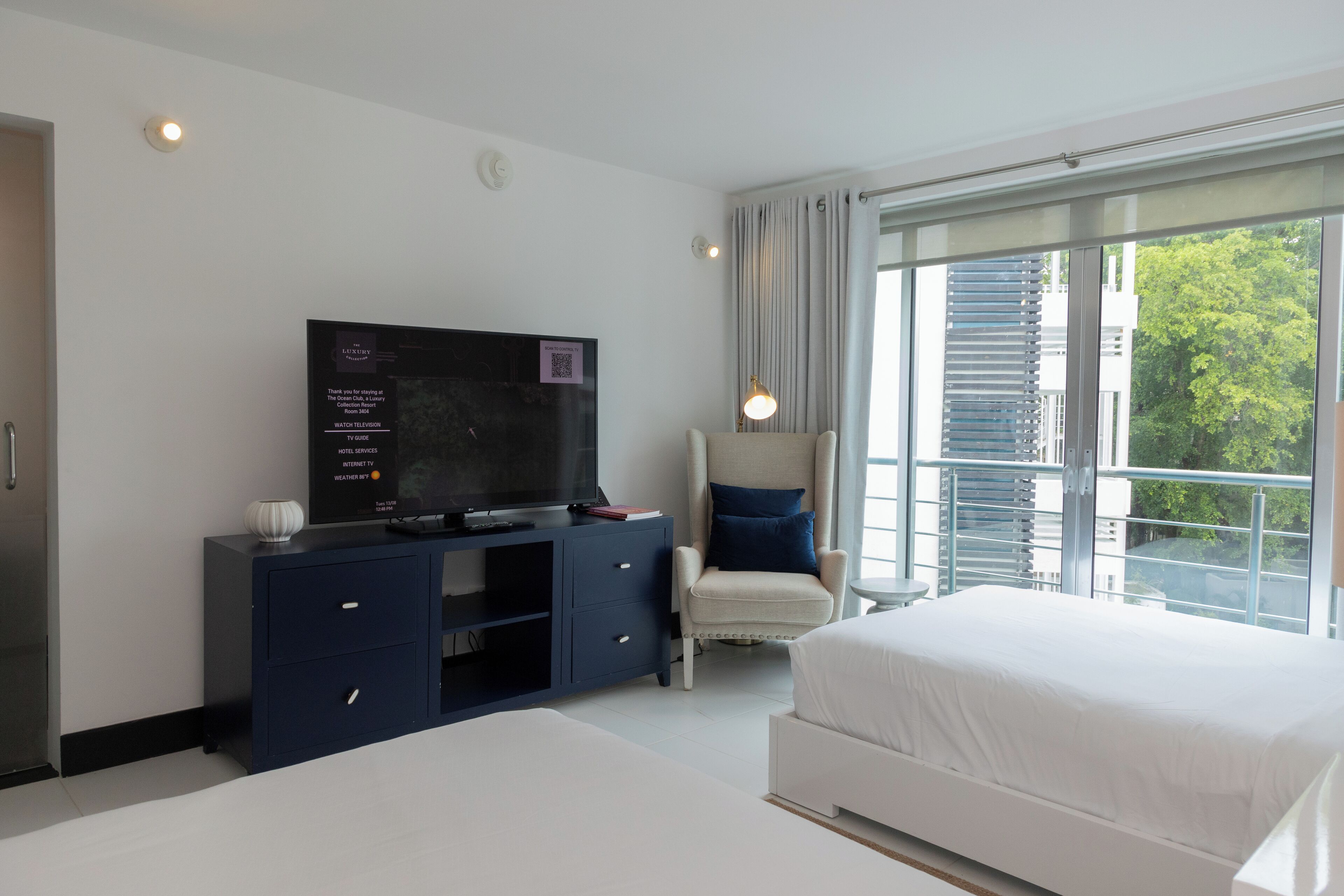 A hotel room with a large flat-screen TV, a comfortable chair, a bed, and a large window offering a view of the city.