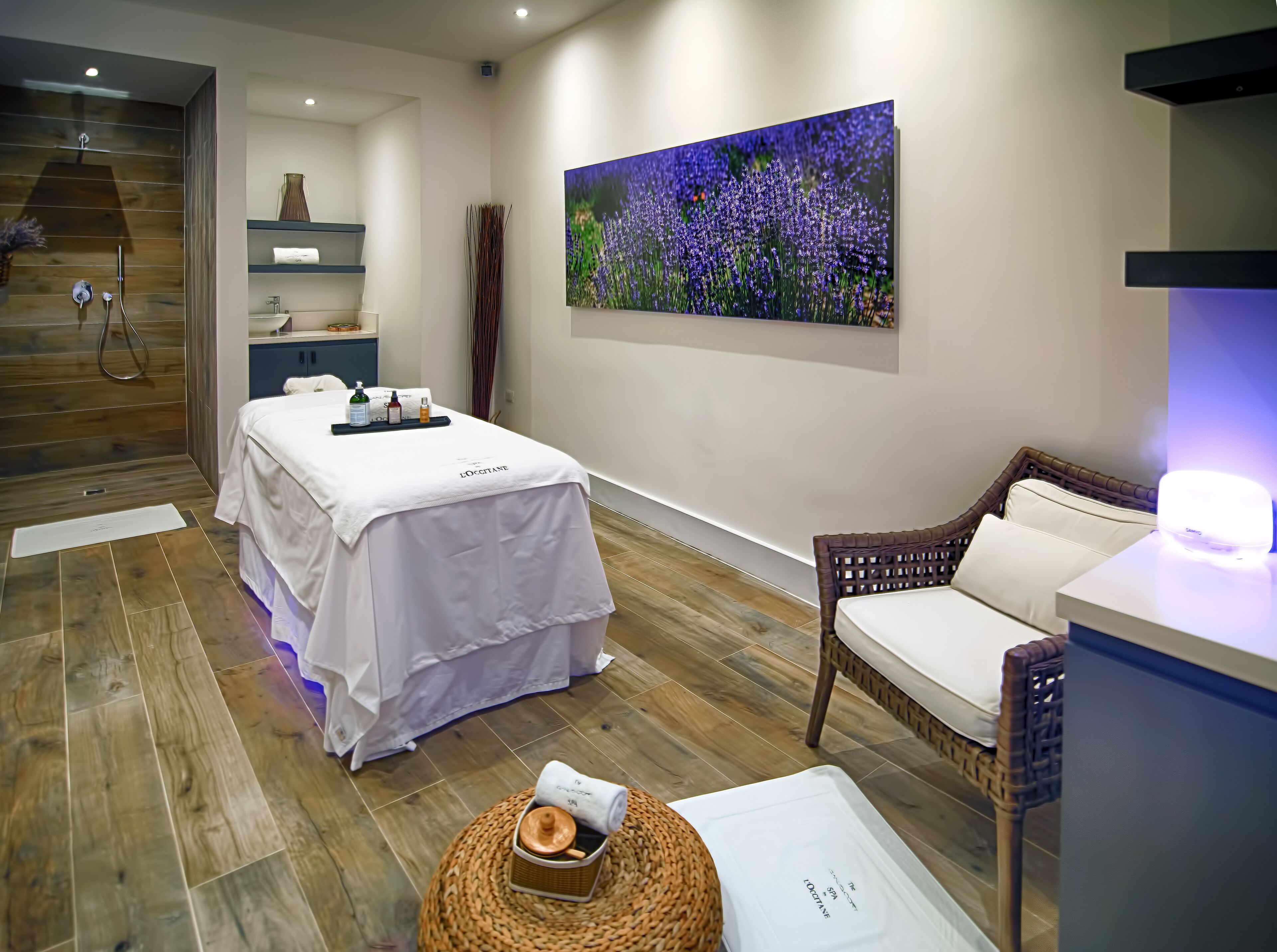 A spa room with a massage table, a wicker chair, a wall-mounted massage chair, and a large painting of flowers.