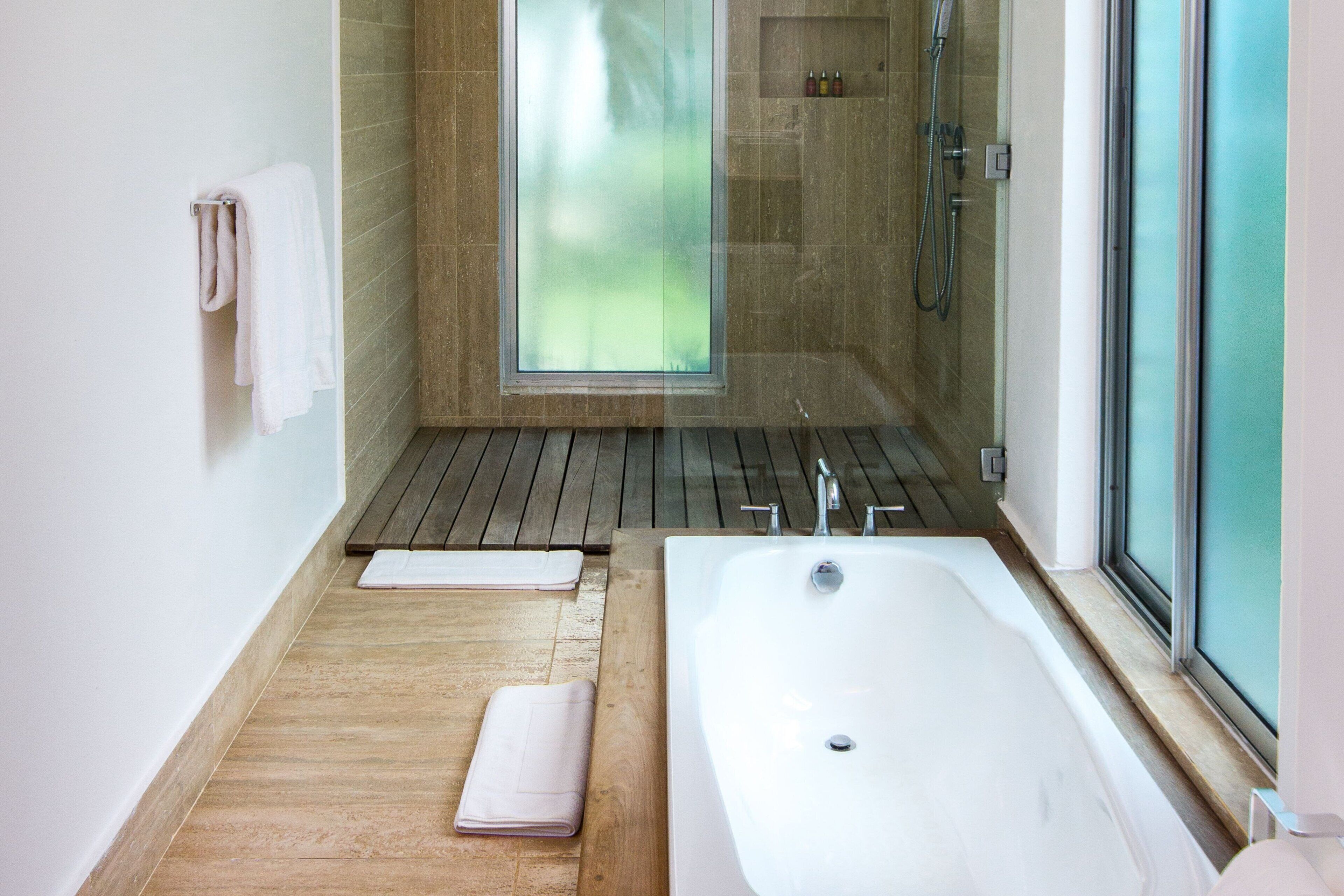 A modern bathroom with a large bathtub, wooden flooring, a glass shower enclosure, and a view of greenery outside.