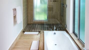 A modern bathroom with a large bathtub, wooden flooring, a glass shower enclosure, and a view of greenery outside.