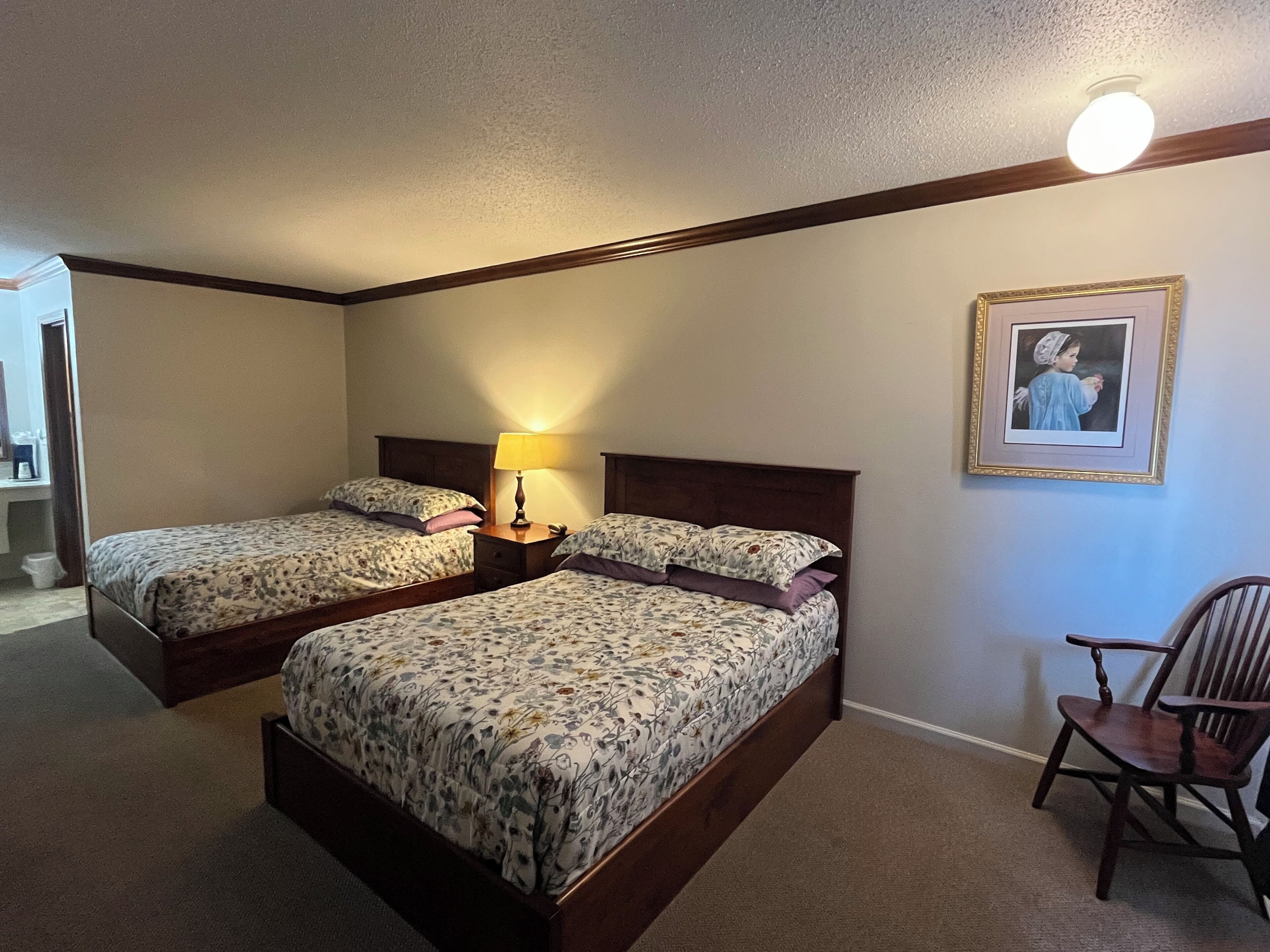 A hotel room with two single beds, a nightstand with a lamp, a framed picture on the wall, and a wooden chair.