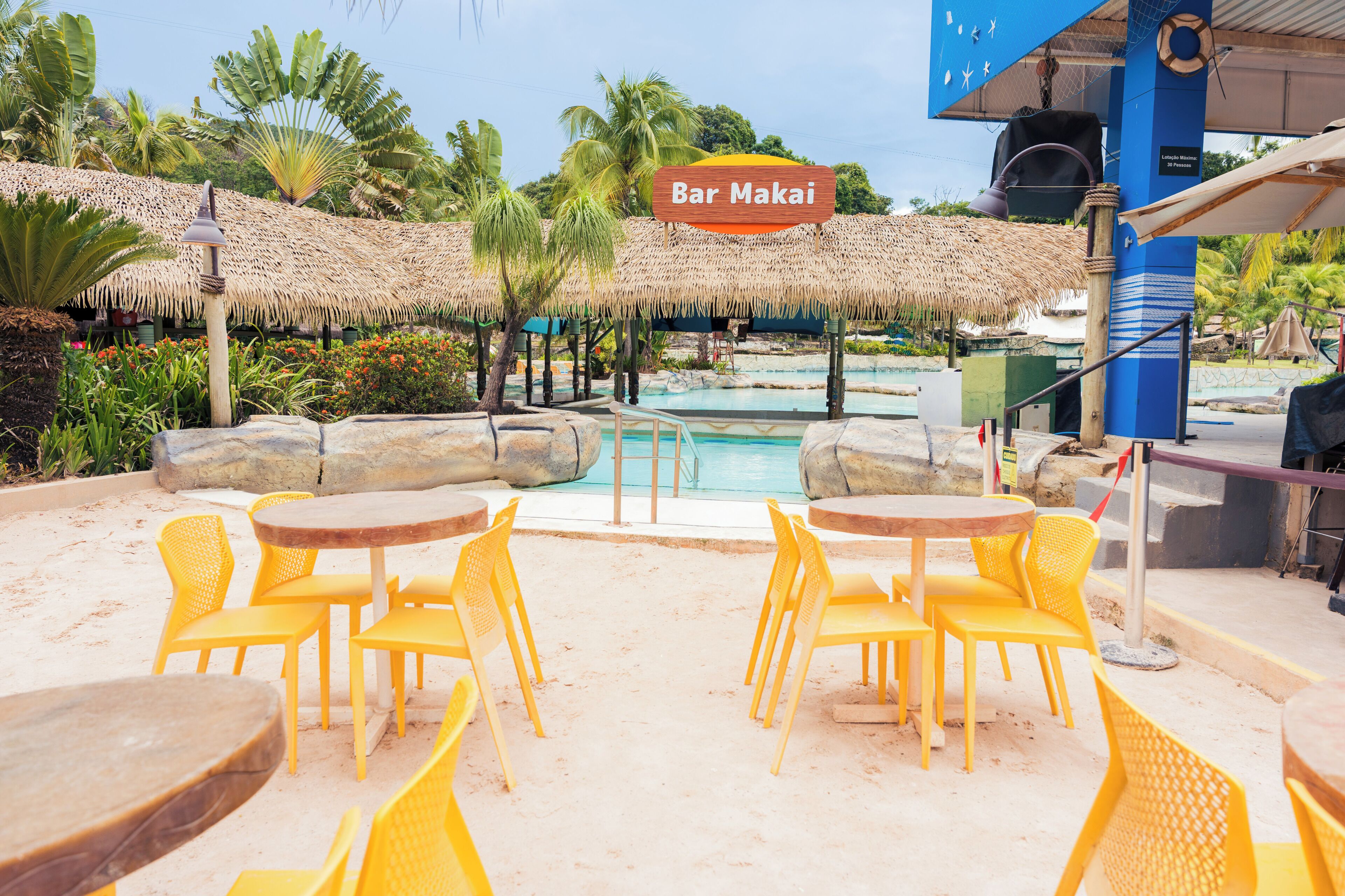 A beachside bar area with yellow chairs and tables, a pool, and thatched umbrellas.