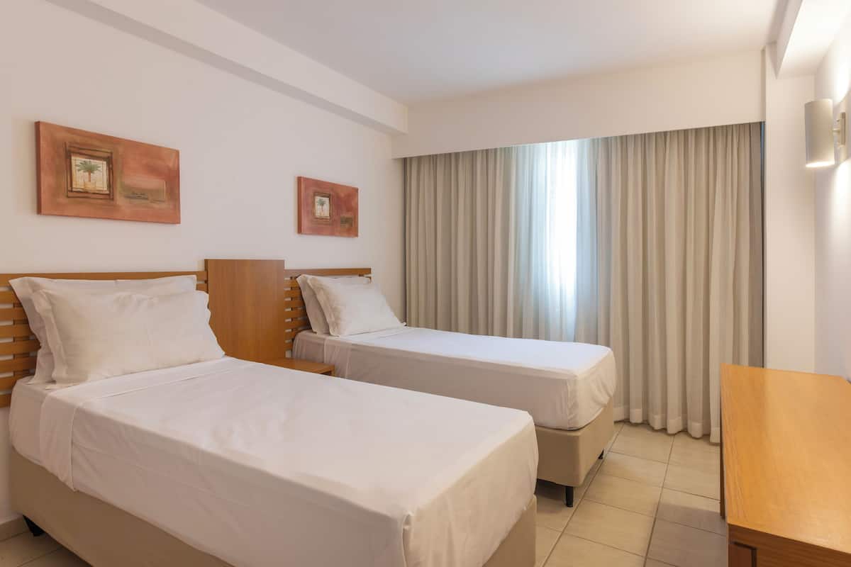 A hotel room with two single beds, a wooden headboard, a small wooden table, and a large window with curtains.
