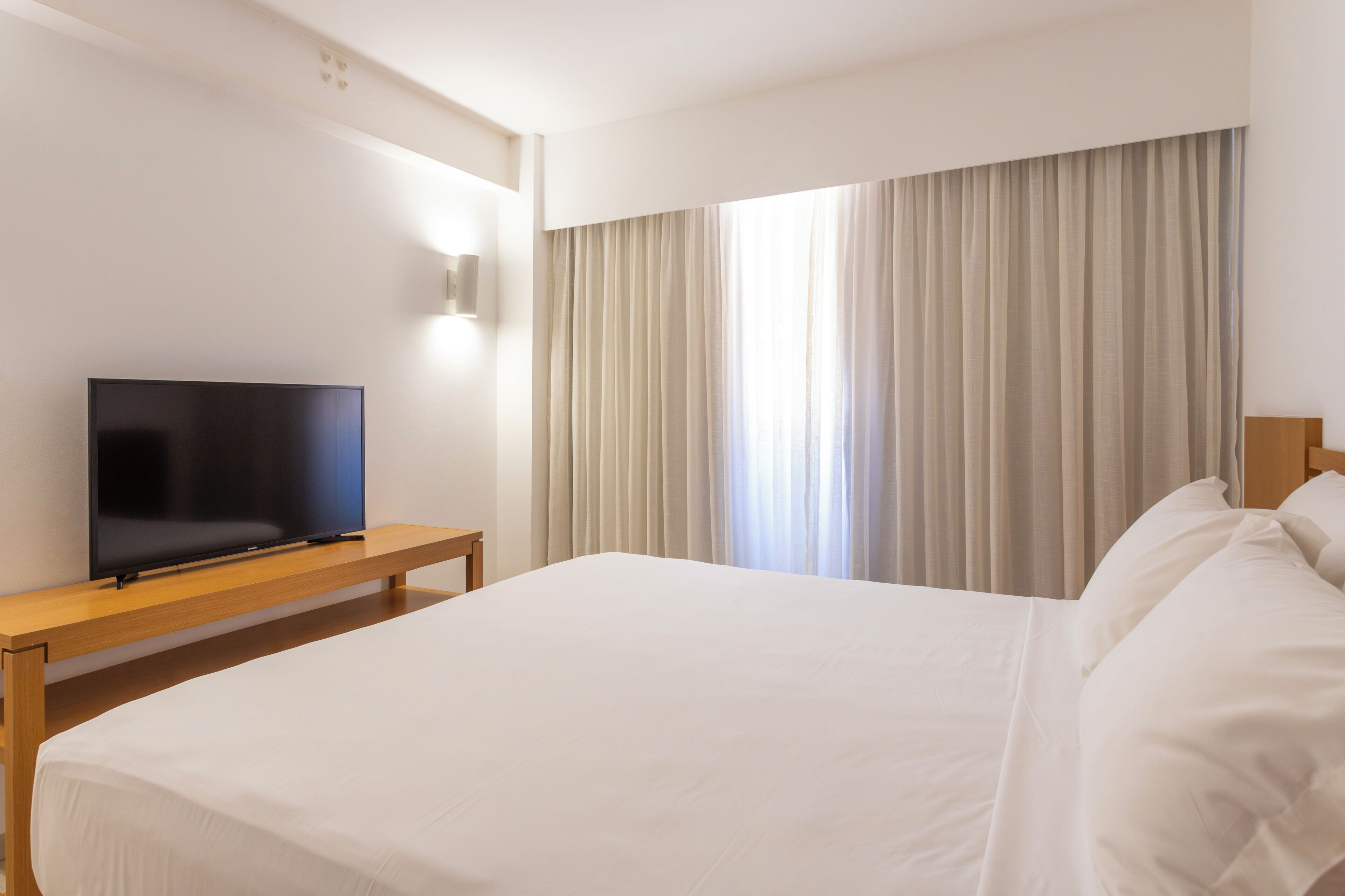 A hotel room with a large bed, a flat-screen TV on a wooden nightstand, and a window with curtains.