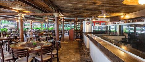 Un restaurante interior con mesas y sillas de madera, una barra con caja registradora y vistas a zonas verdes al exterior.