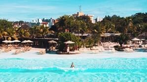 A beach with clear turquoise water, a person swimming, and a resort area with umbrellas and lounge chairs. - Rio Quente Resorts - Hotel Luupi (Rio Quente)