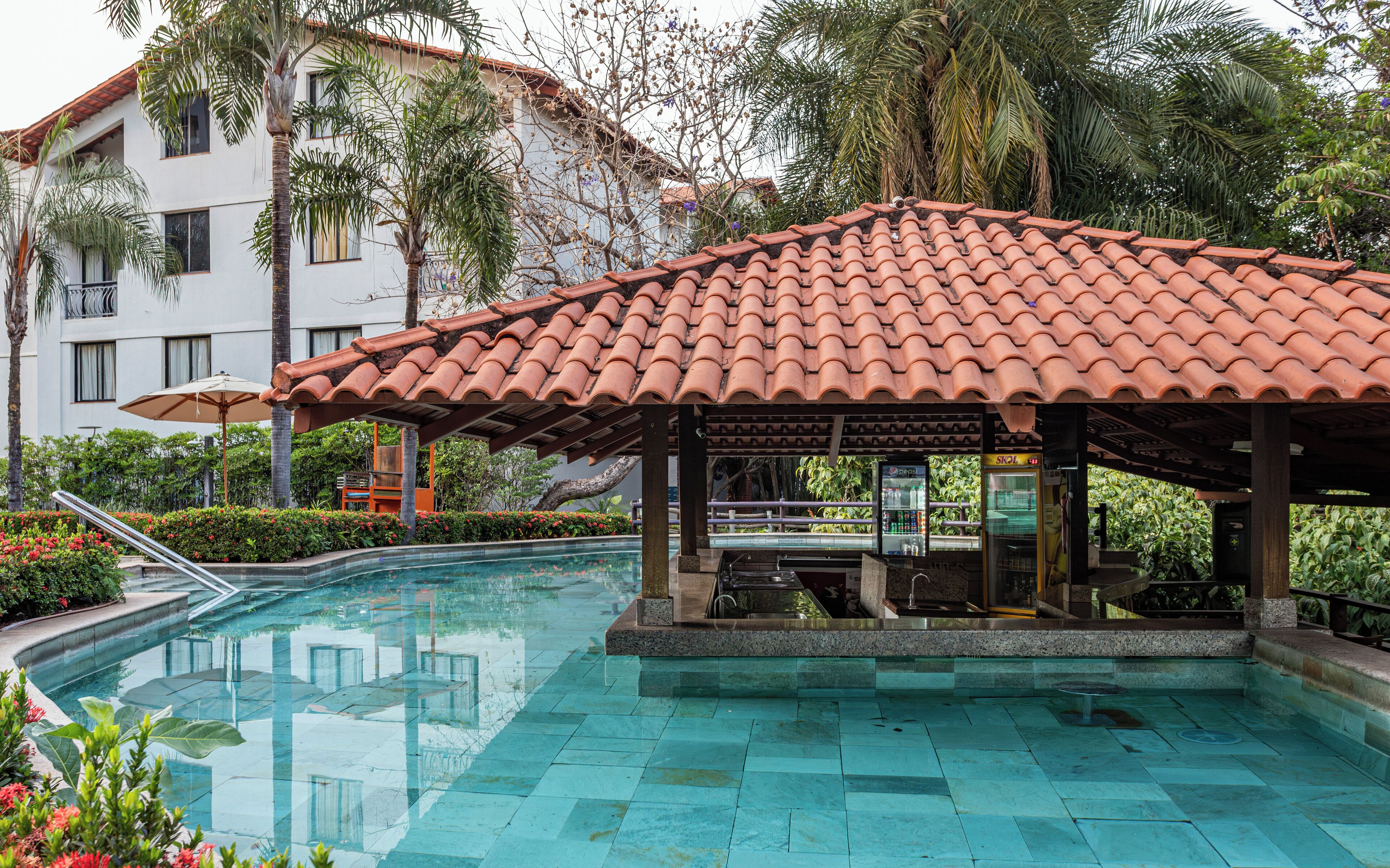 A swimming pool with a tiled roof and a bar area, surrounded by lush greenery and a building with a tiled roof.
