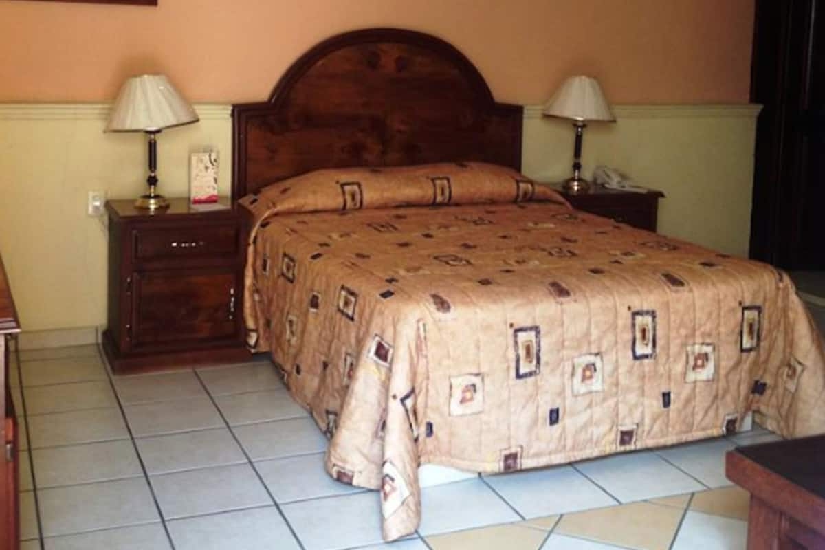 Un dormitorio con una cama de madera, dos mesitas de noche con lámparas, un cuadro en la pared y suelo de baldosas.
