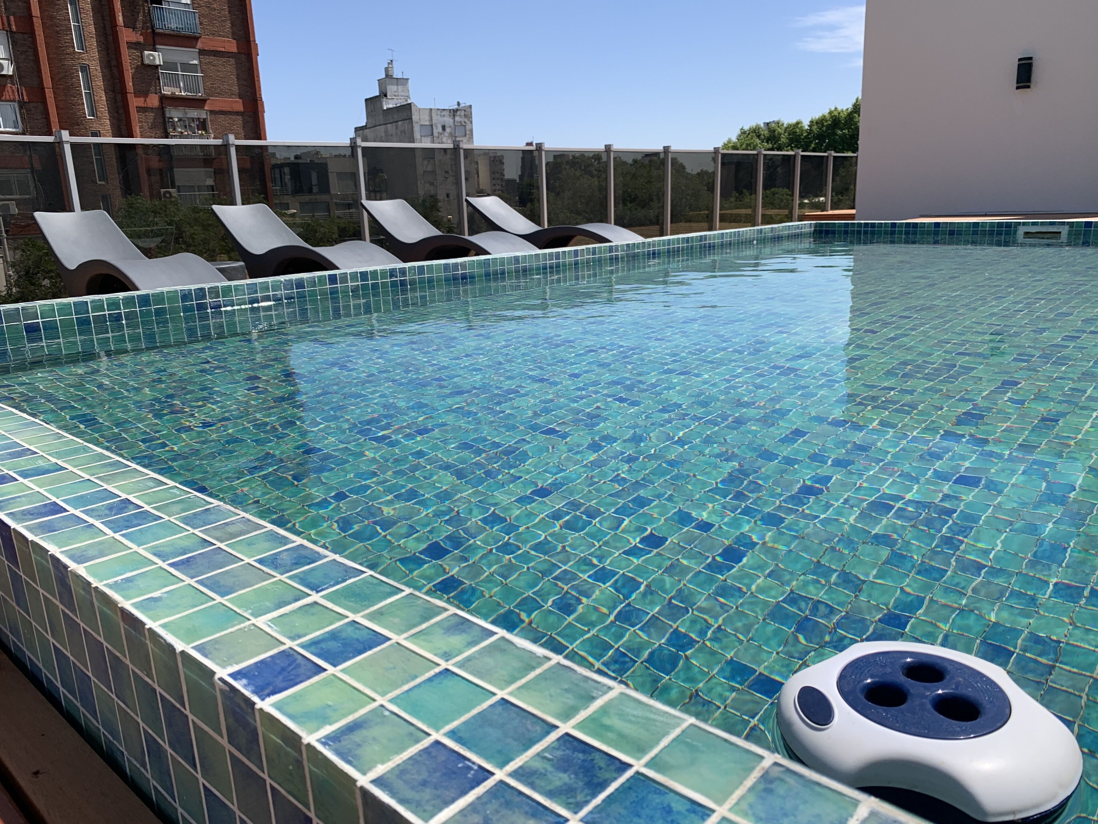 A swimming pool with blue mosaic tiles, lounge chairs, and a view of buildings in the background.