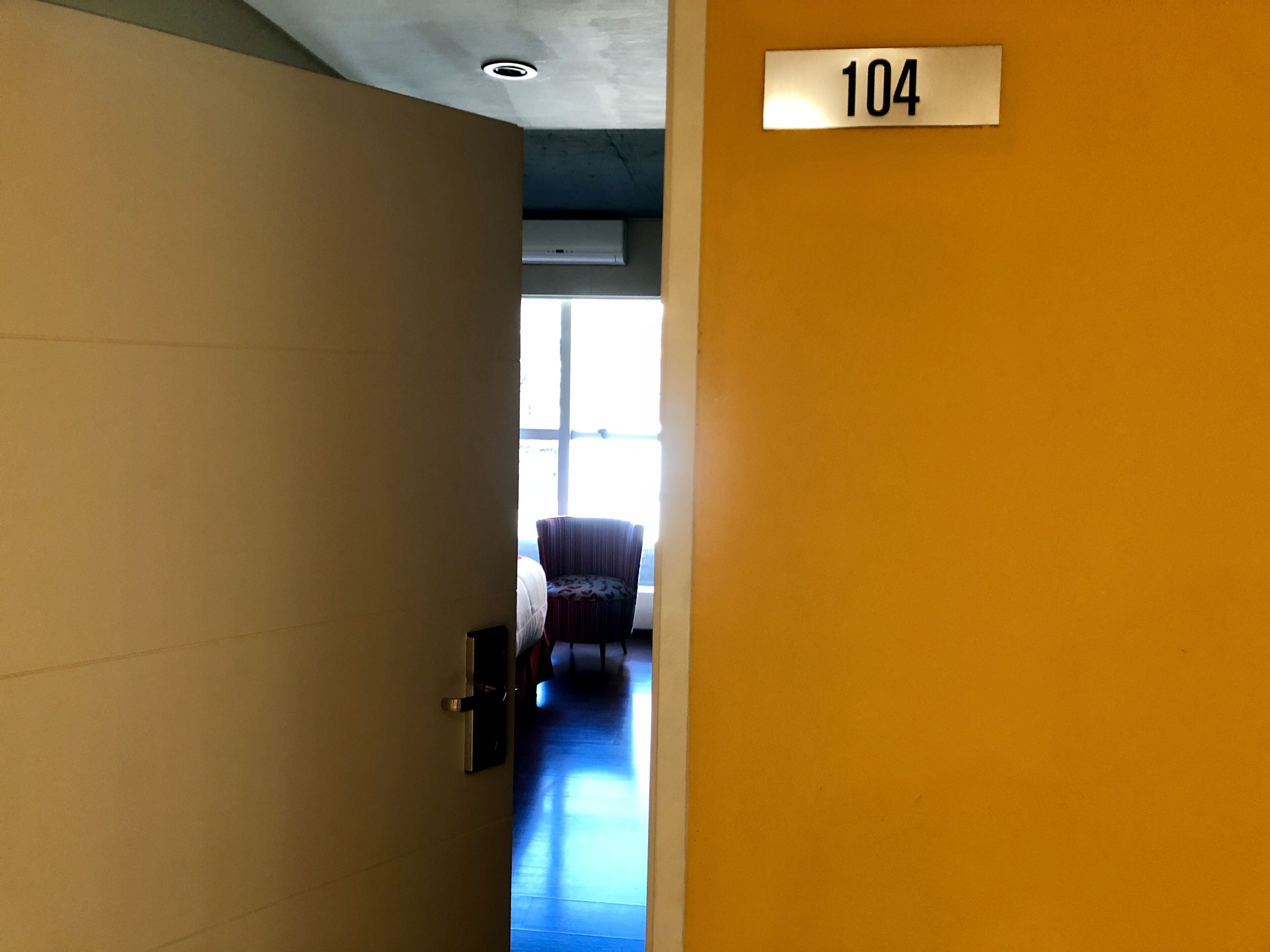 Un couloir avec une porte portant l’inscription « 104 », qui donne accès à une pièce contenant un lit et une chaise.