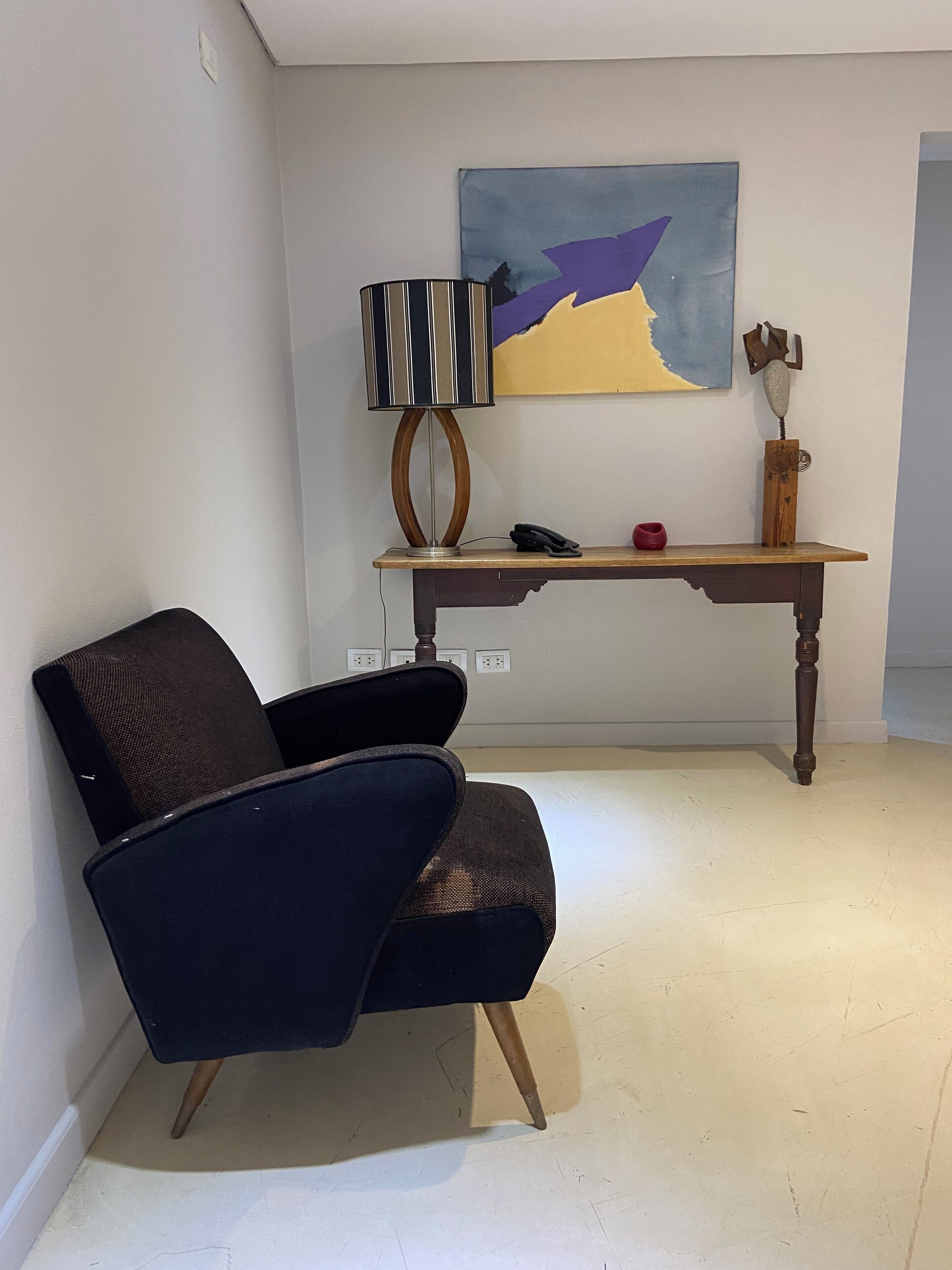A room with a dark blue armchair, a wooden table with a lamp, a painting, and a small sculpture.