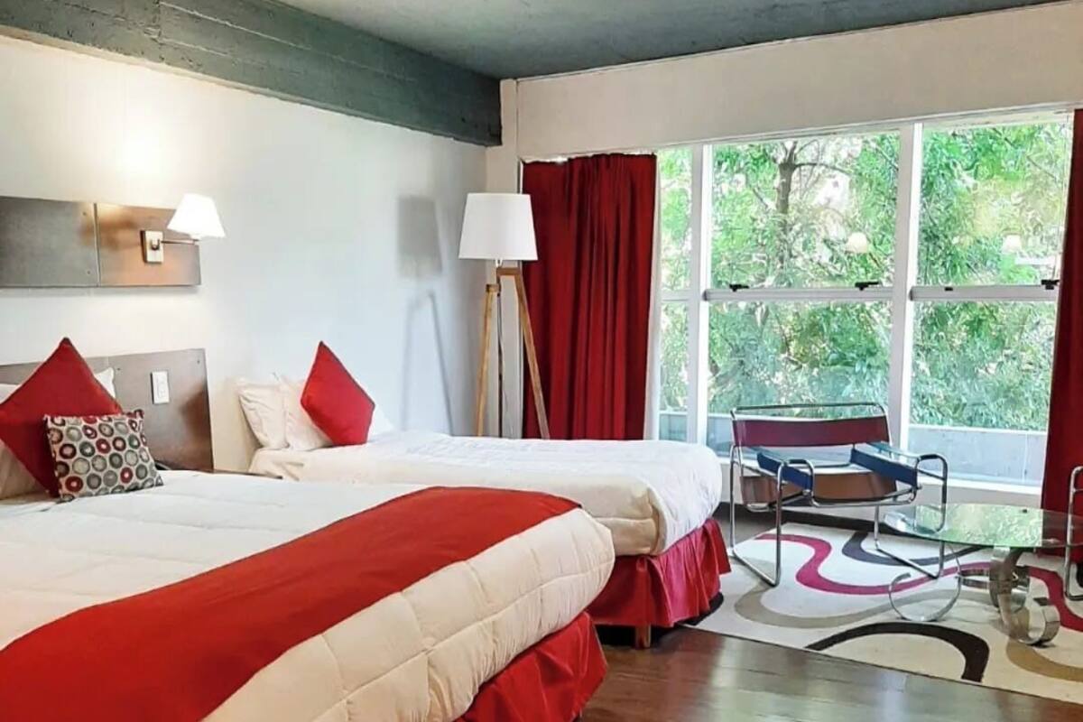 A hotel room with two beds, a large window with red curtains, a standing lamp, and a view of greenery outside.