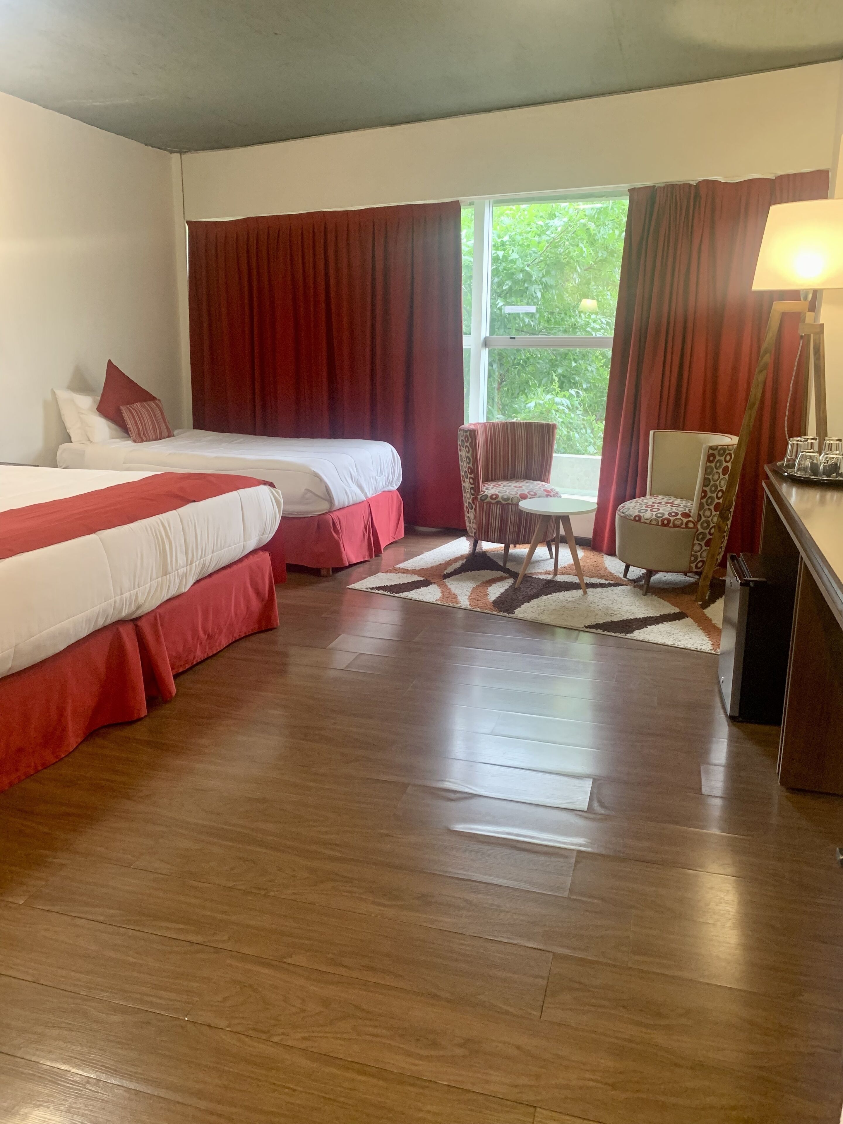 Habitación de hotel con dos camas, una mesita, una silla y una ventana con cortinas.