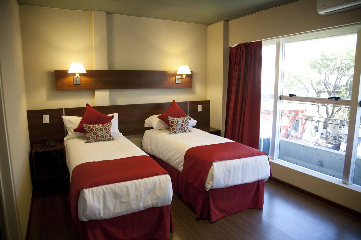 A hotel room with two beds, each with a red bedspread and matching pillows, a nightstand with a lamp, and a window with a view of the outside.