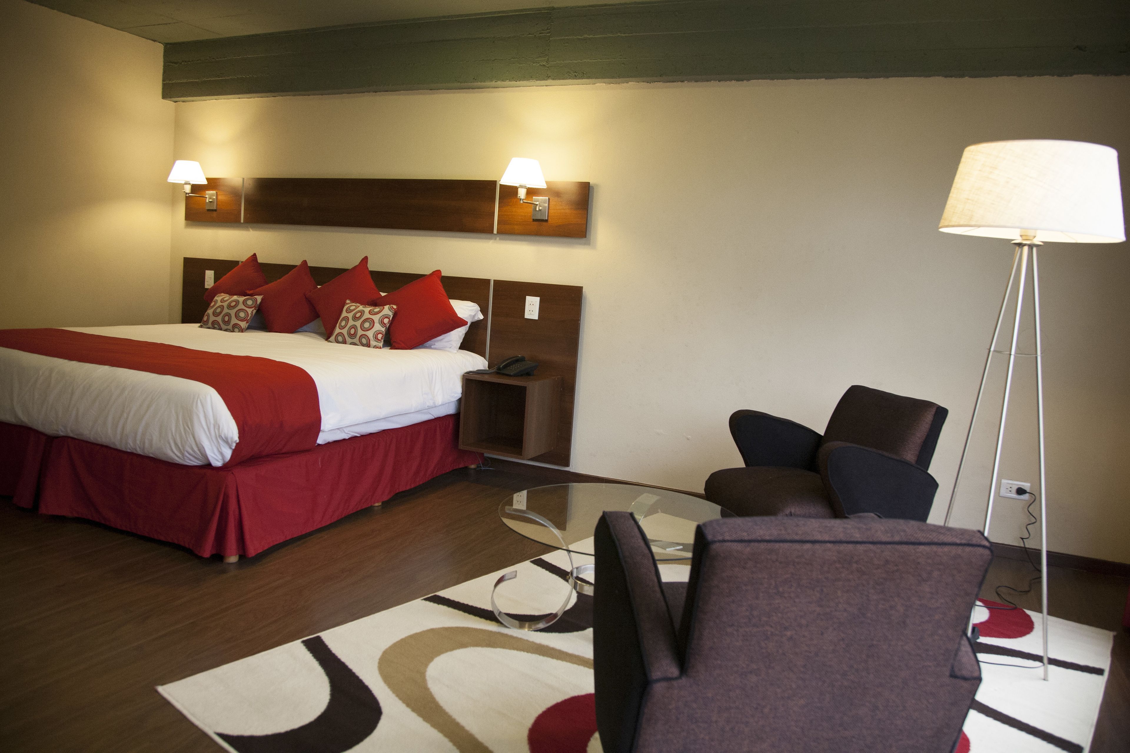 Habitación de hotel con cama, mesita de noche, lámpara y una mesa de vidrio con sillas.