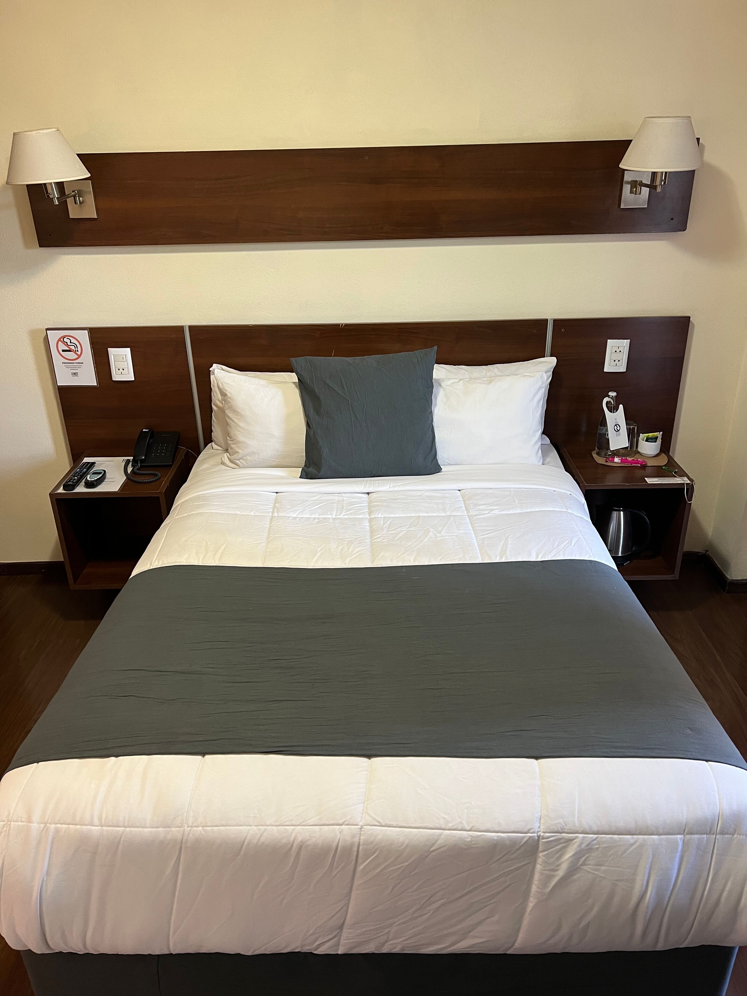 A hotel room with a large bed, two bedside tables, a nightstand, a wall-mounted lamp, a telephone, and a sign indicating no smoking.