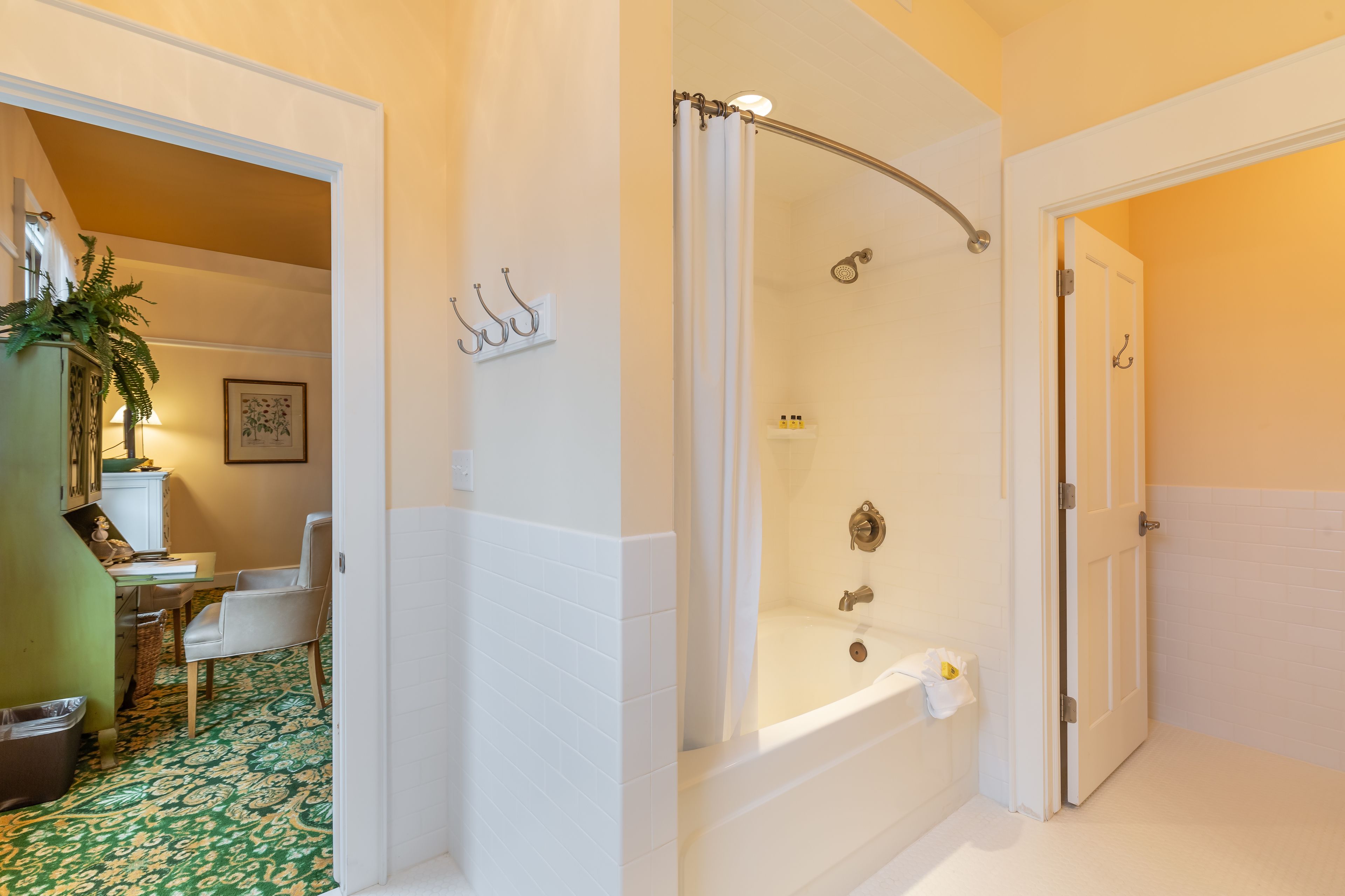Un baño con bañera, ducha e inodoro. Hay una puerta que da a otra habitación, una silla y una planta.