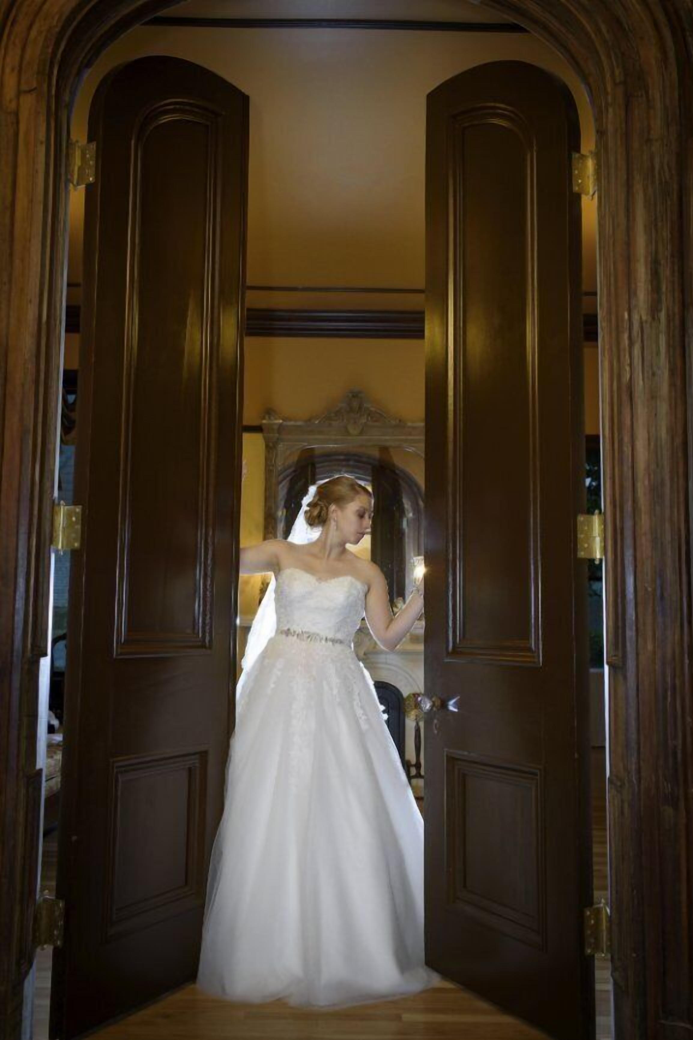 Una mujer con un vestido de novia parada en un umbral con puertas de madera y un espejo.