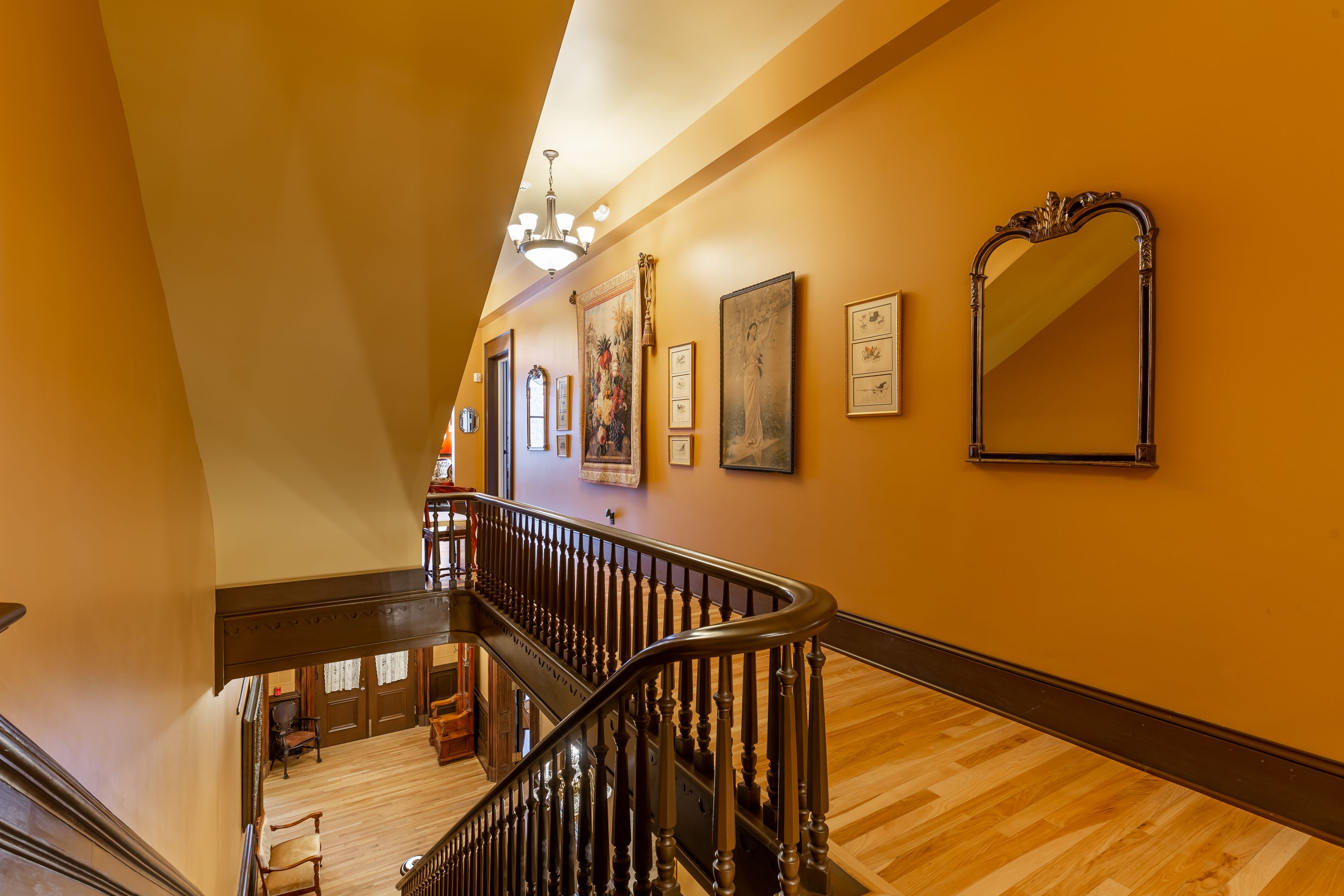 Una imponente escalera con barandas de madera y un candelabro, que conduce a un espacio en el segundo piso con obras de arte enmarcadas y un espejo grande.