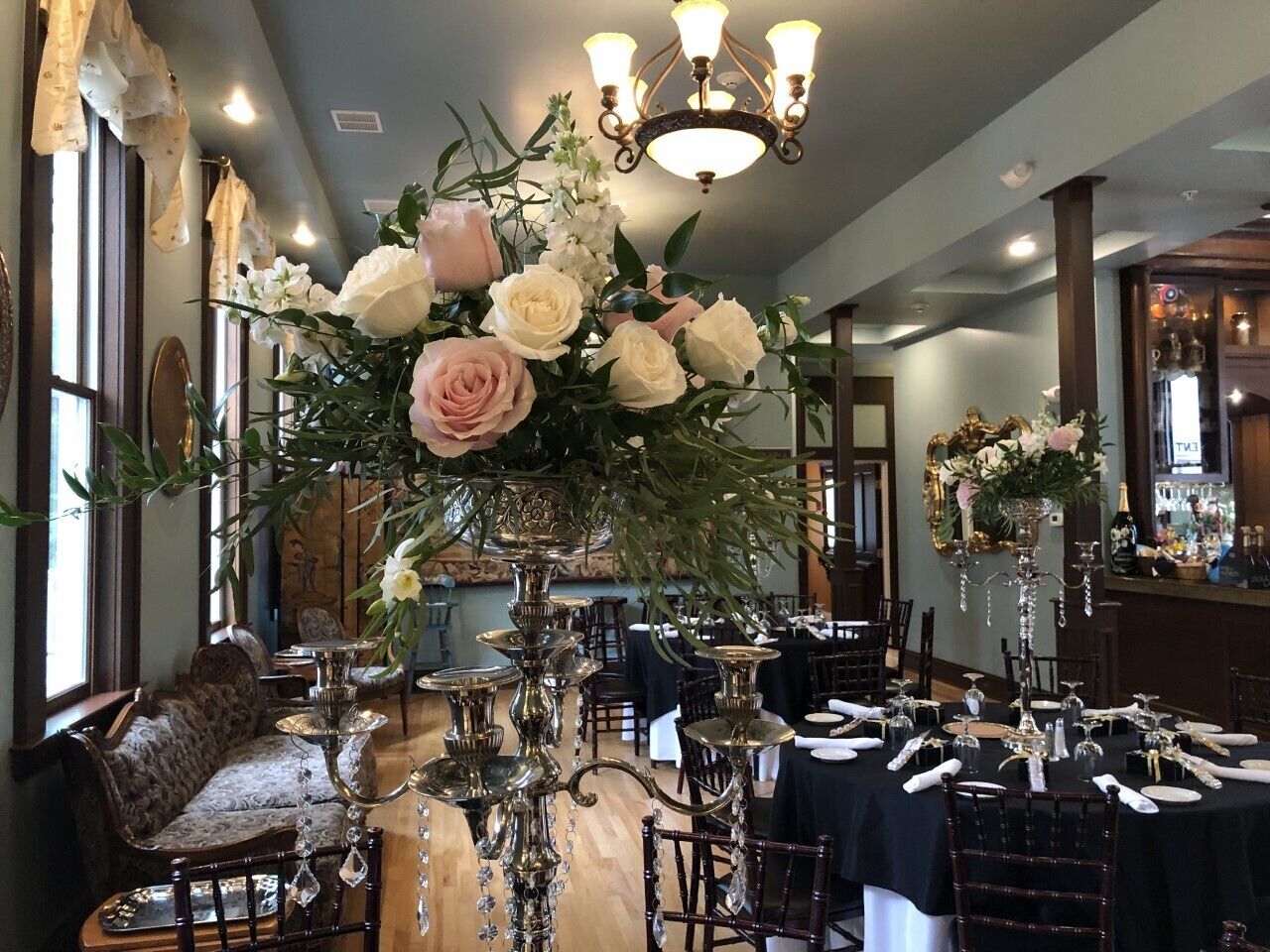 Uma sala de jantar formal com mesas postas para uma refeição, um grande arranjo floral e um candelabro.