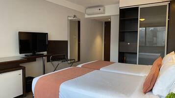Quarto de hotel com duas camas, televisão, escrivaninha, espelho e guarda-roupa.