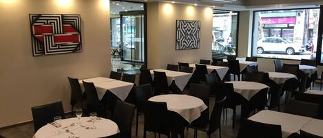 A restaurant interior with round tables, black chairs, and white tablecloths. There are abstract art pieces on the walls and a view of the street outside.