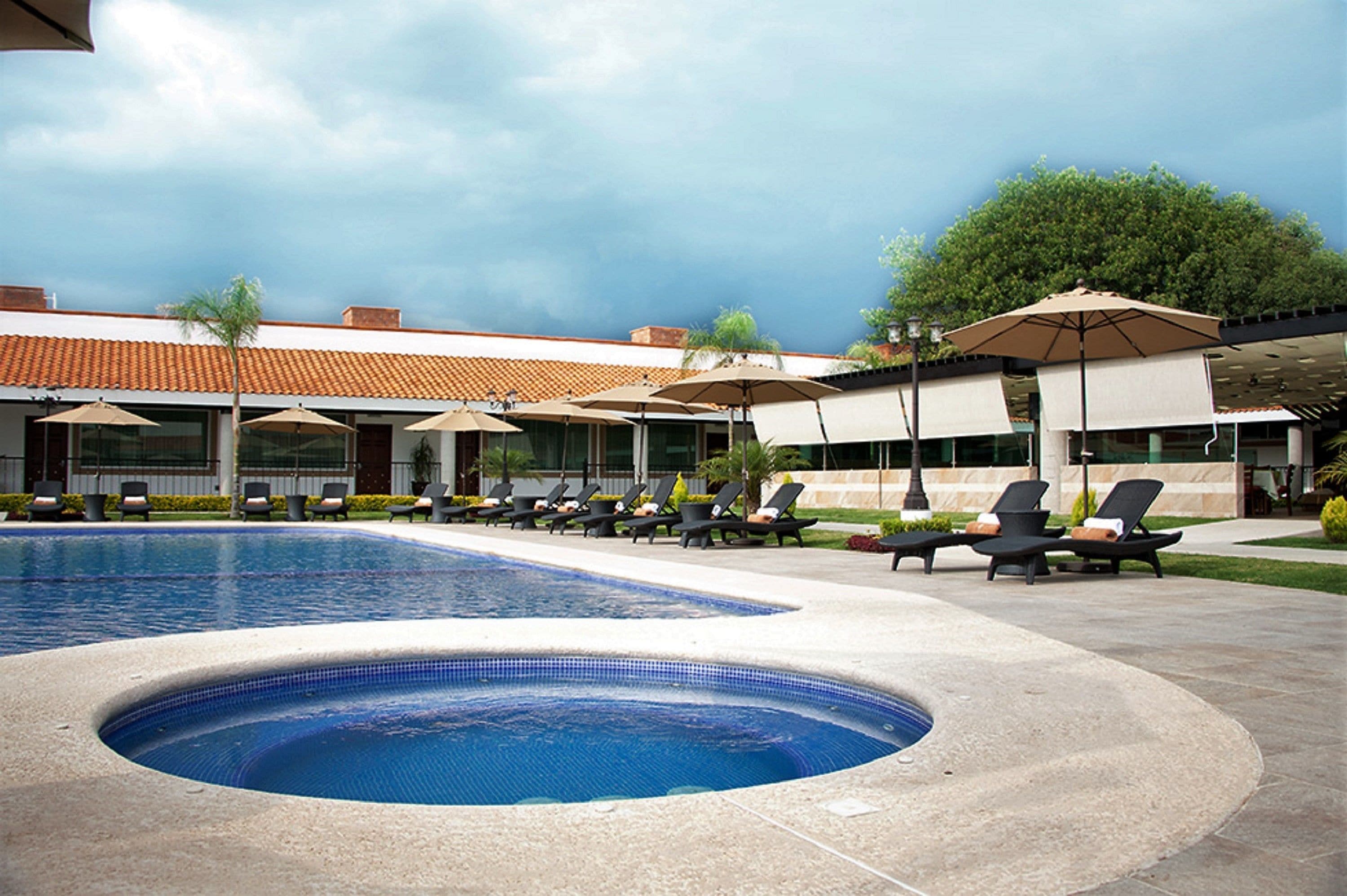 A swimming pool with lounge chairs and umbrellas, surrounded by a building with a tiled roof and a tree.