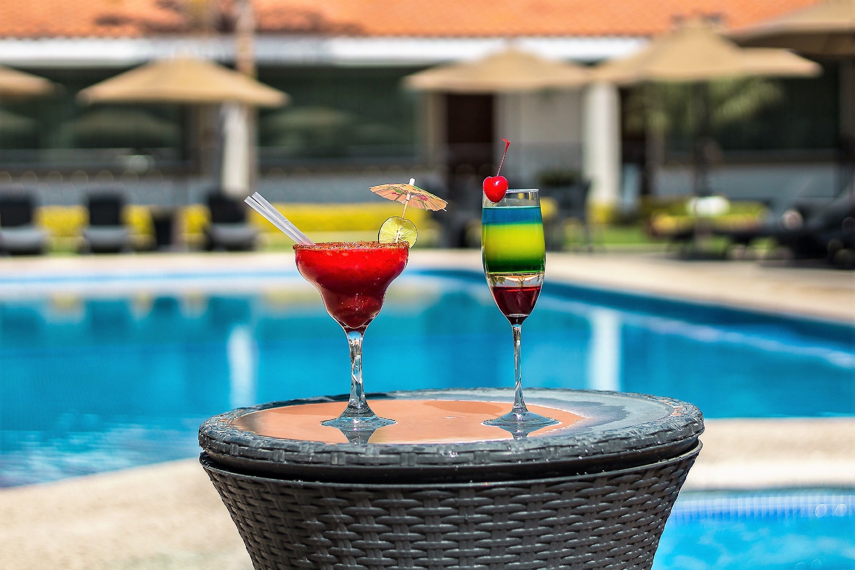 Dos cócteles sobre una mesa de mimbre junto a una piscina con sillas de descanso y sombrillas al fondo.