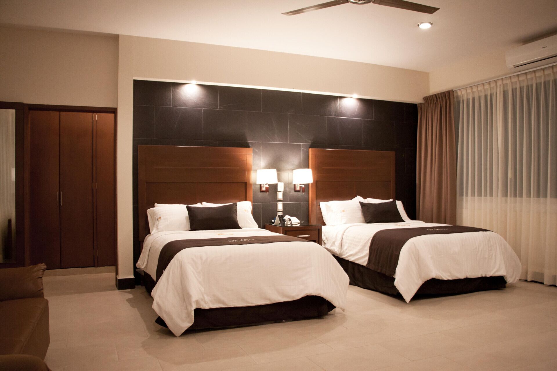 Habitación de hotel con dos camas, cabecero de madera, lámparas de pared, ventilador de techo y un ventanal con cortinas.