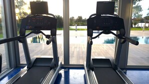 Two exercise machines with digital displays, positioned in front of a large window overlooking a swimming pool. - Faros de Carrasco Apartments (Paso de Carrasco)
