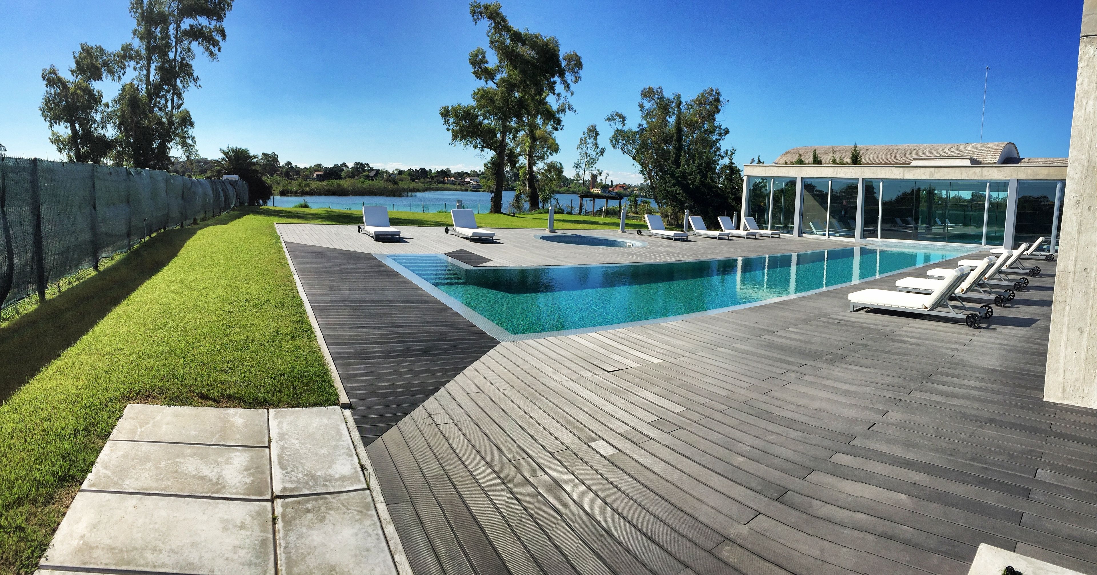 Un área de piscina moderna al aire libre con cubierta de madera, sillas de descanso y vista a un lago.