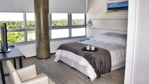 A modern hotel room with a large bed, a desk with a computer, a chair, and a view of a lake through large windows. - Faros de Carrasco Apartments (Paso de Carrasco)