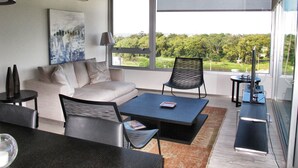 A modern living room with a sofa, a coffee table, a TV stand, and a view of a green landscape through large windows. - Faros de Carrasco Apartments (Paso de Carrasco)