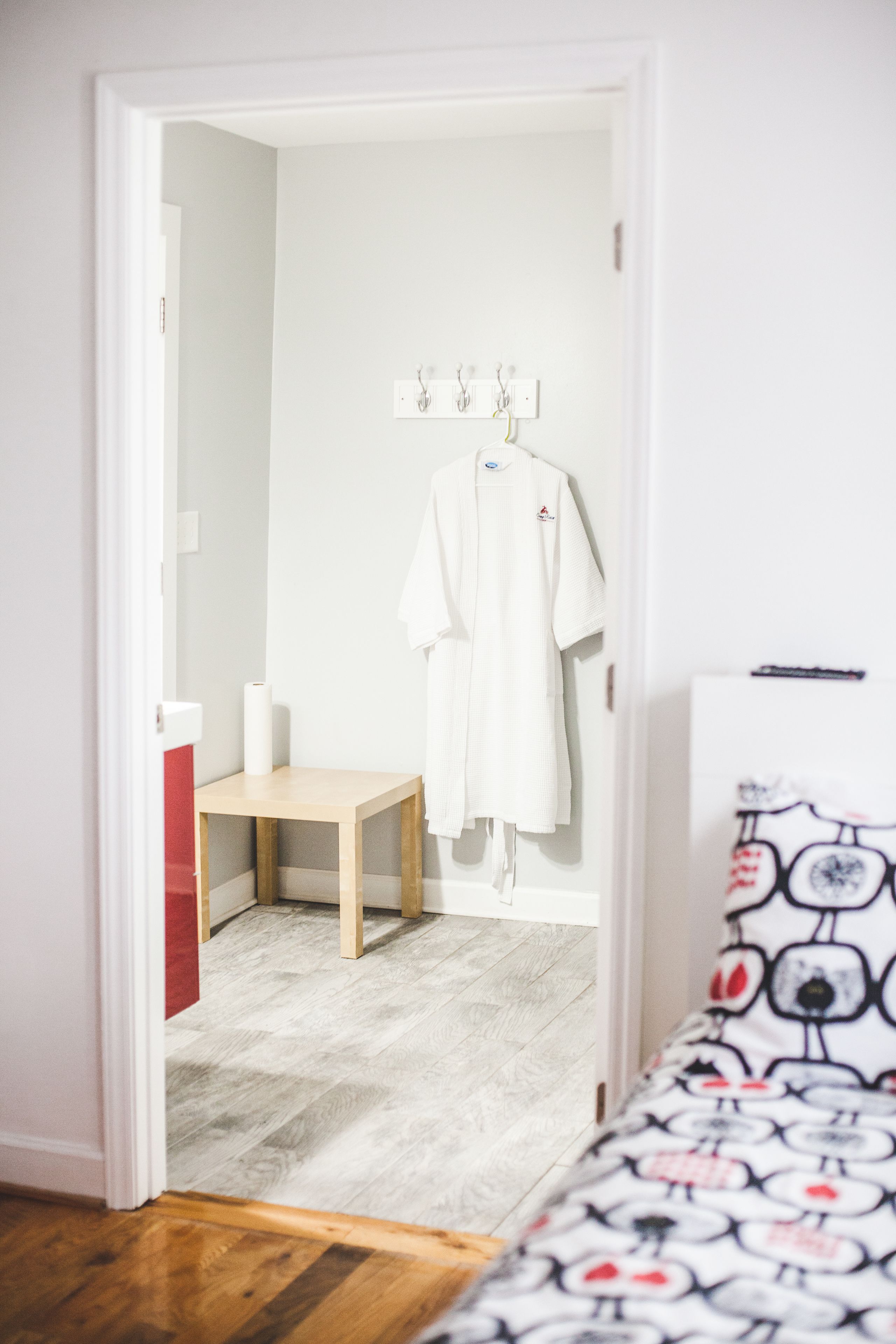 Ein Schlafzimmer mit einem Bett, einem Nachttisch aus Holz, einem kleinen Tisch und einem weißen Bademantel, der an einem Haken hängt.