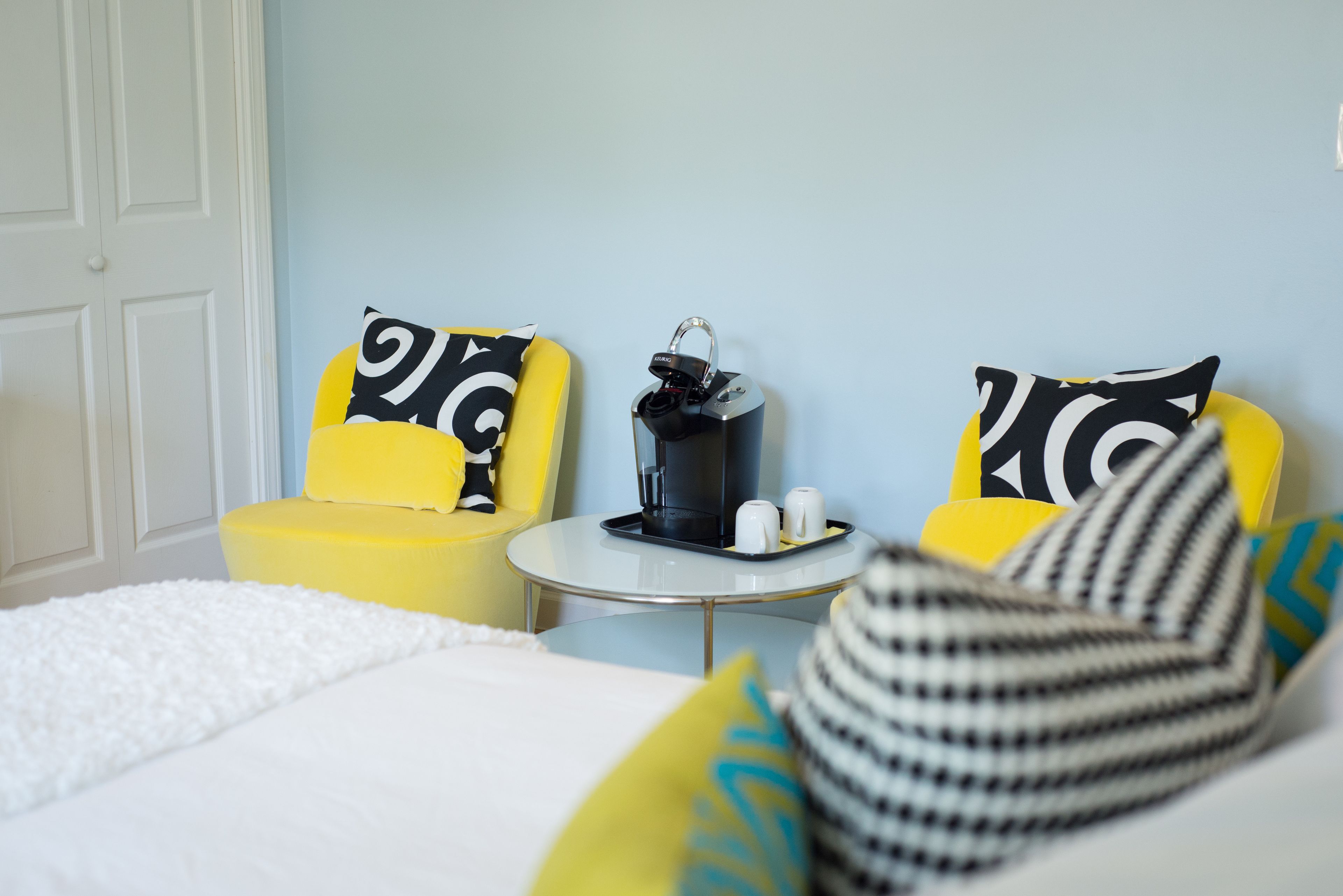 Una habitación de hotel con cama, dos sillas amarillas con cojines de diseño blanco y negro, una mesita redonda con cafetera y tazas, y una puerta blanca.