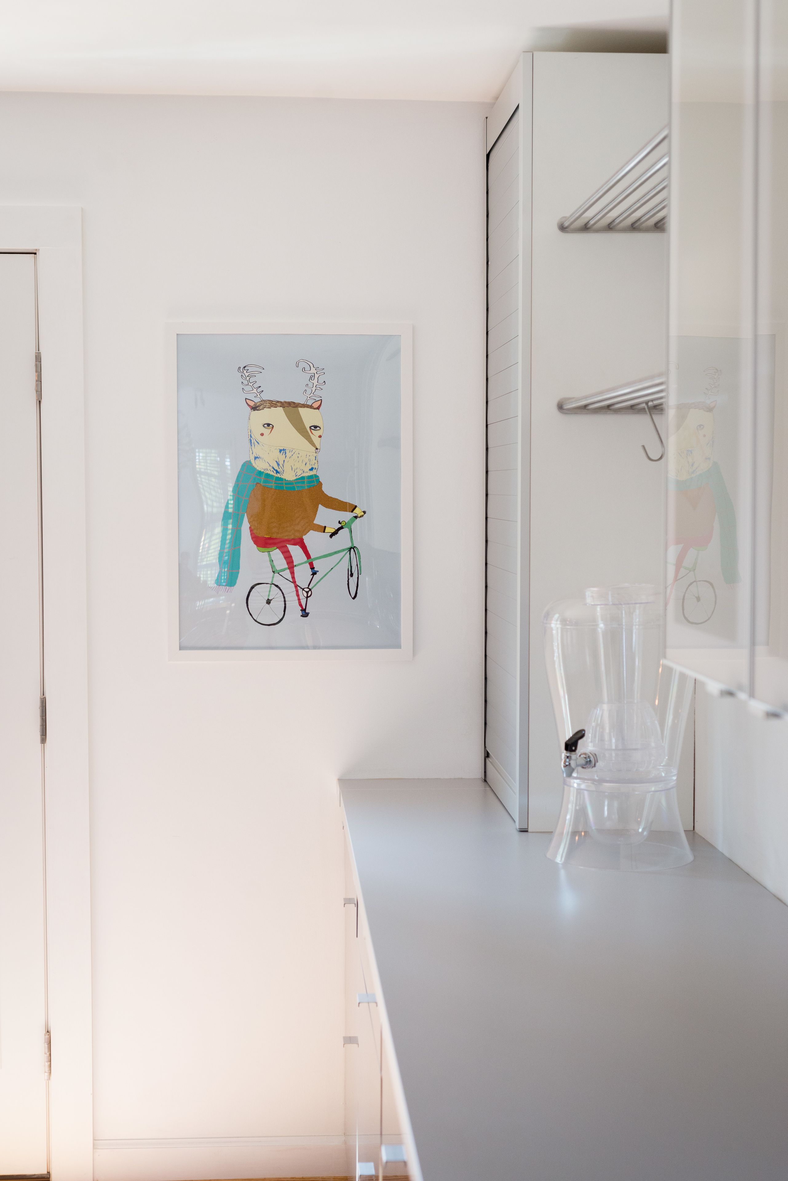 A room with a white wall, a framed artwork of a character on a bicycle, a white cabinet, and a clear plastic container on the counter.