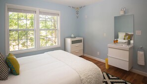 A bedroom with a large window, a bed with white bedding, a nightstand, a dresser, a mirror, and a bathroom with a sink and toilet.