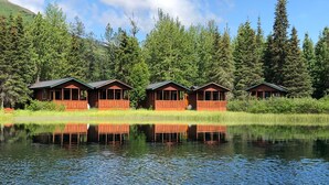 A row of wooden cabins with a lake in the foreground and a forested hillside in the background. - Summit Lake Lodge (Moose Pass)