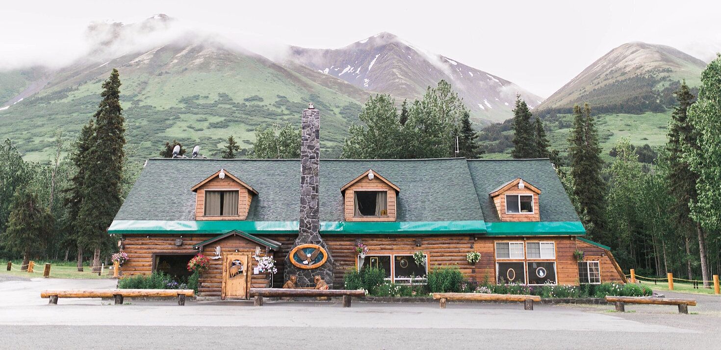 A rustic log cabin with a stone chimney, wooden windows, and a green roof.