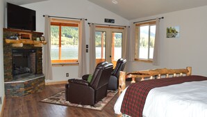 A bedroom with a fireplace, a bed, a TV, and a view of a lake. - Summit Lake Lodge (Moose Pass)