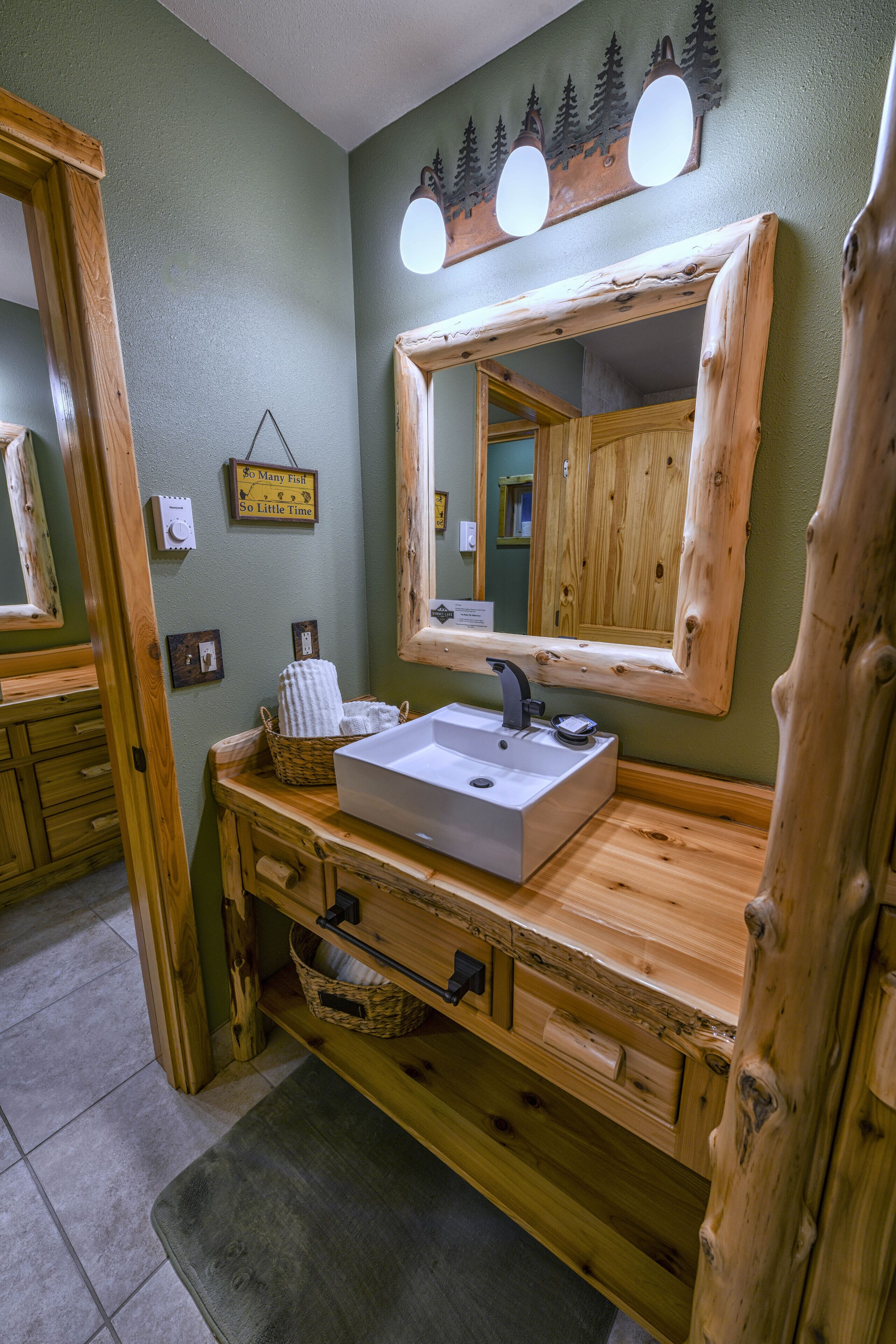 A bathroom with a wooden vanity, a white sink, and a large mirror. There are lights above the mirror and a small sign on the wall.
