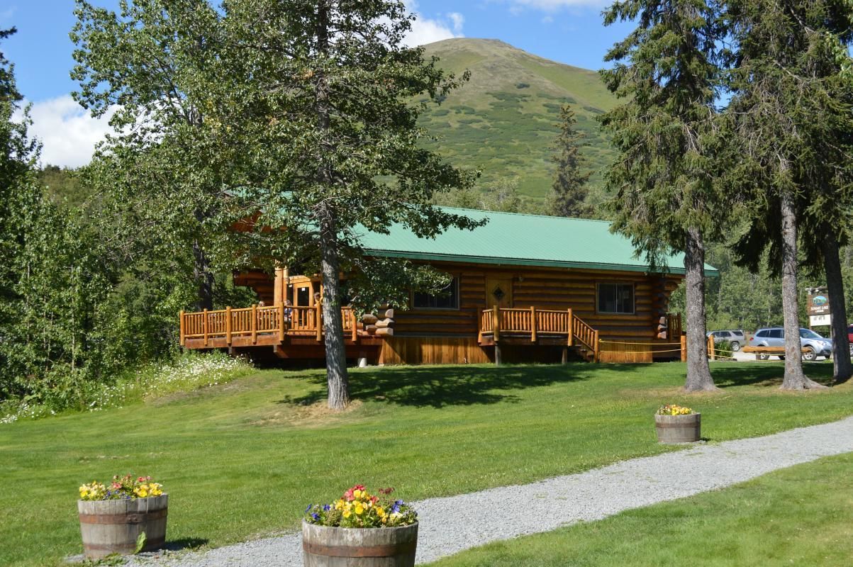 A wooden cabin with a green roof, surrounded by trees and a mountain in the background.