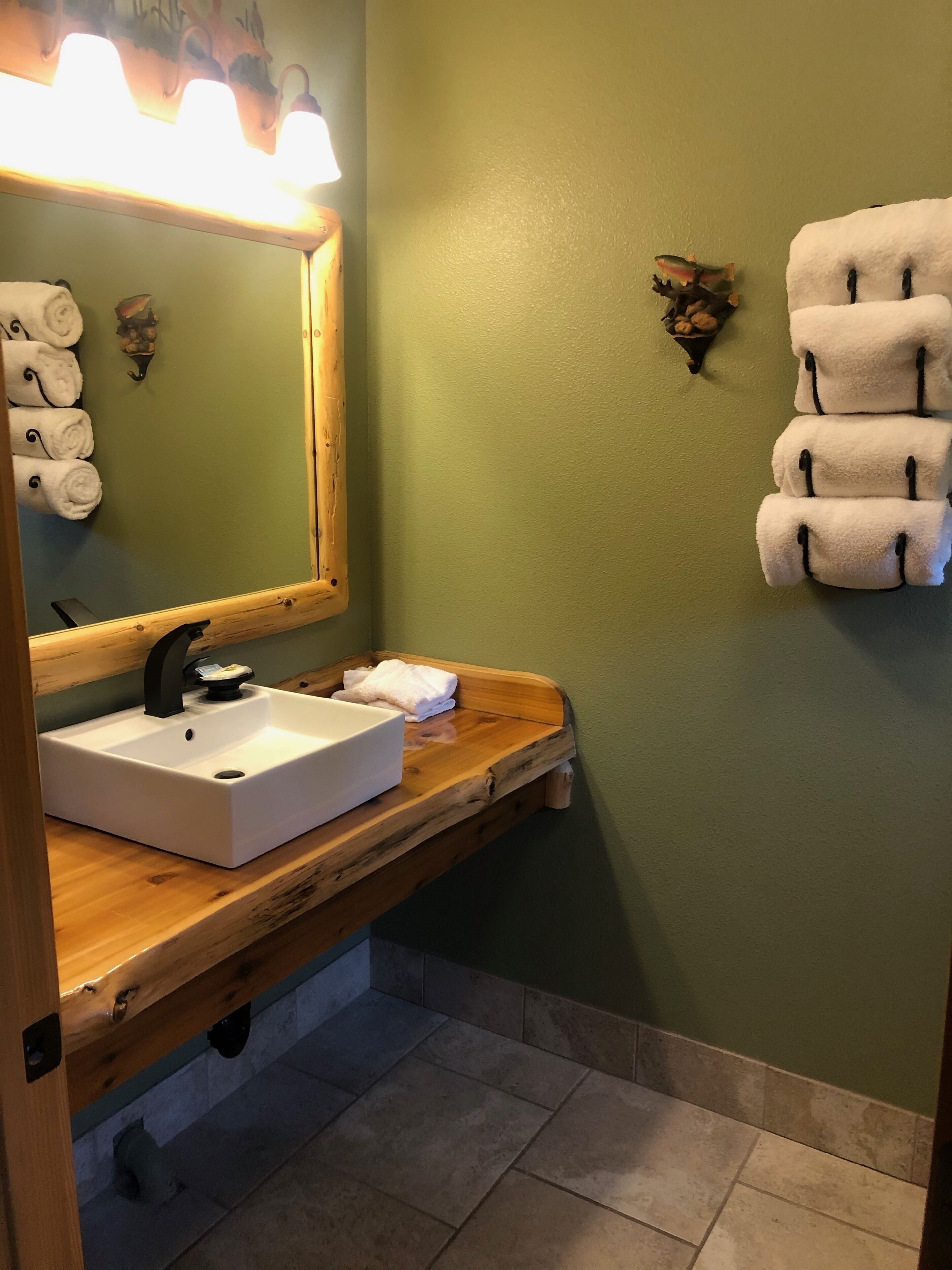 A bathroom with a wooden vanity, a white sink, and a mirror with a light above it. There are towels hanging on the wall and a small decorative item on the wall.