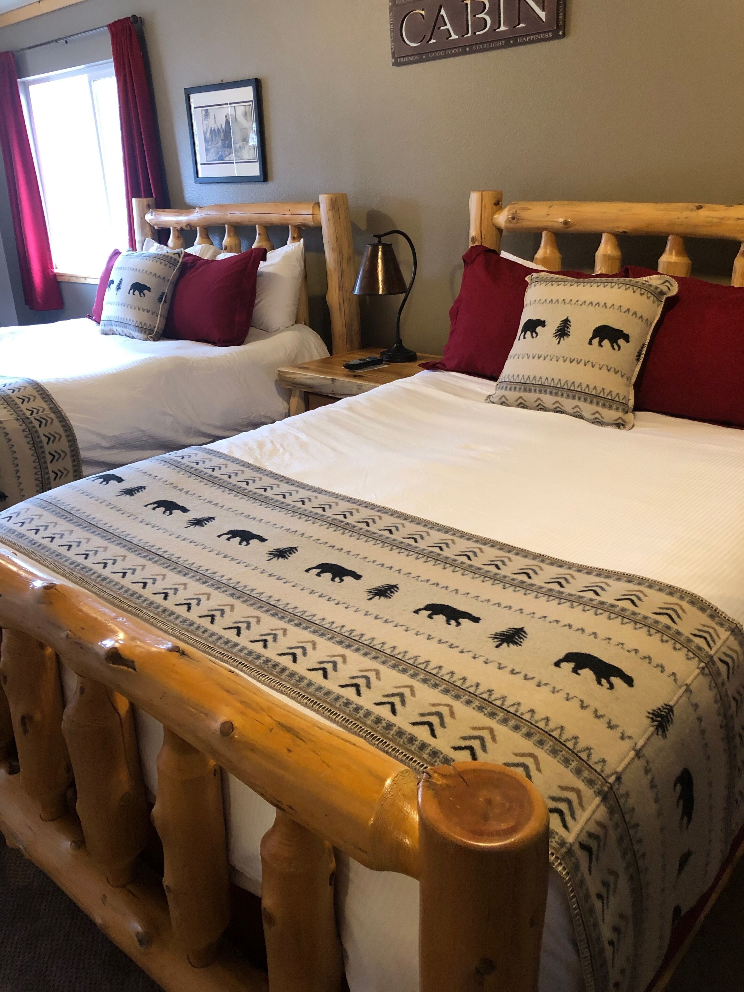 Une chambre de style cabane avec deux lits, chacun doté d’une tête de lit en bois et d’oreillers rouges. Un tableau encadré est accroché au mur, et une pancarte indique « CABANE ».