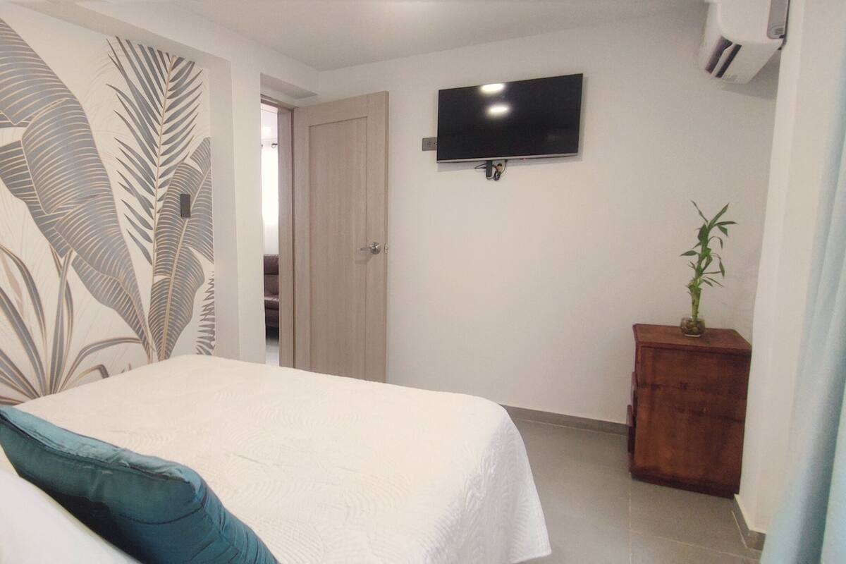 Un dormitorio con cama, almohadas, un televisor montado en la pared y un pequeño aparador de madera con una planta.